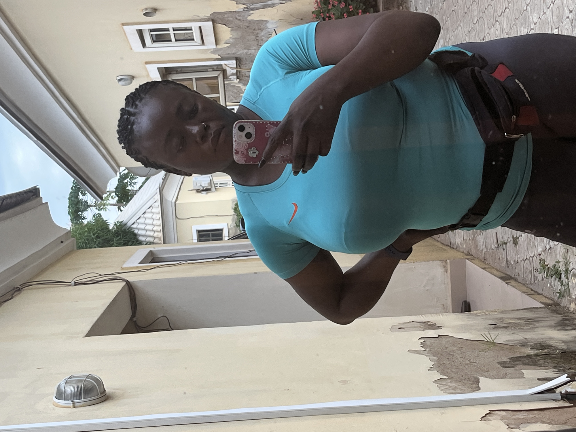 A dark-skinned woman with braided hair takes a selfie in an alleyway in Abuja, Nigeria. She is wearing a turquoise t-shirt, dark leggings, and a black fanny pack around her waist. She is holding a phone with a floral case in her right hand. The alleyway has weathered yellow buildings on either side, with peeling paint and exposed brick. There is a light fixture on the left wall and electrical wires running along the building. The ground is paved with cobblestones.