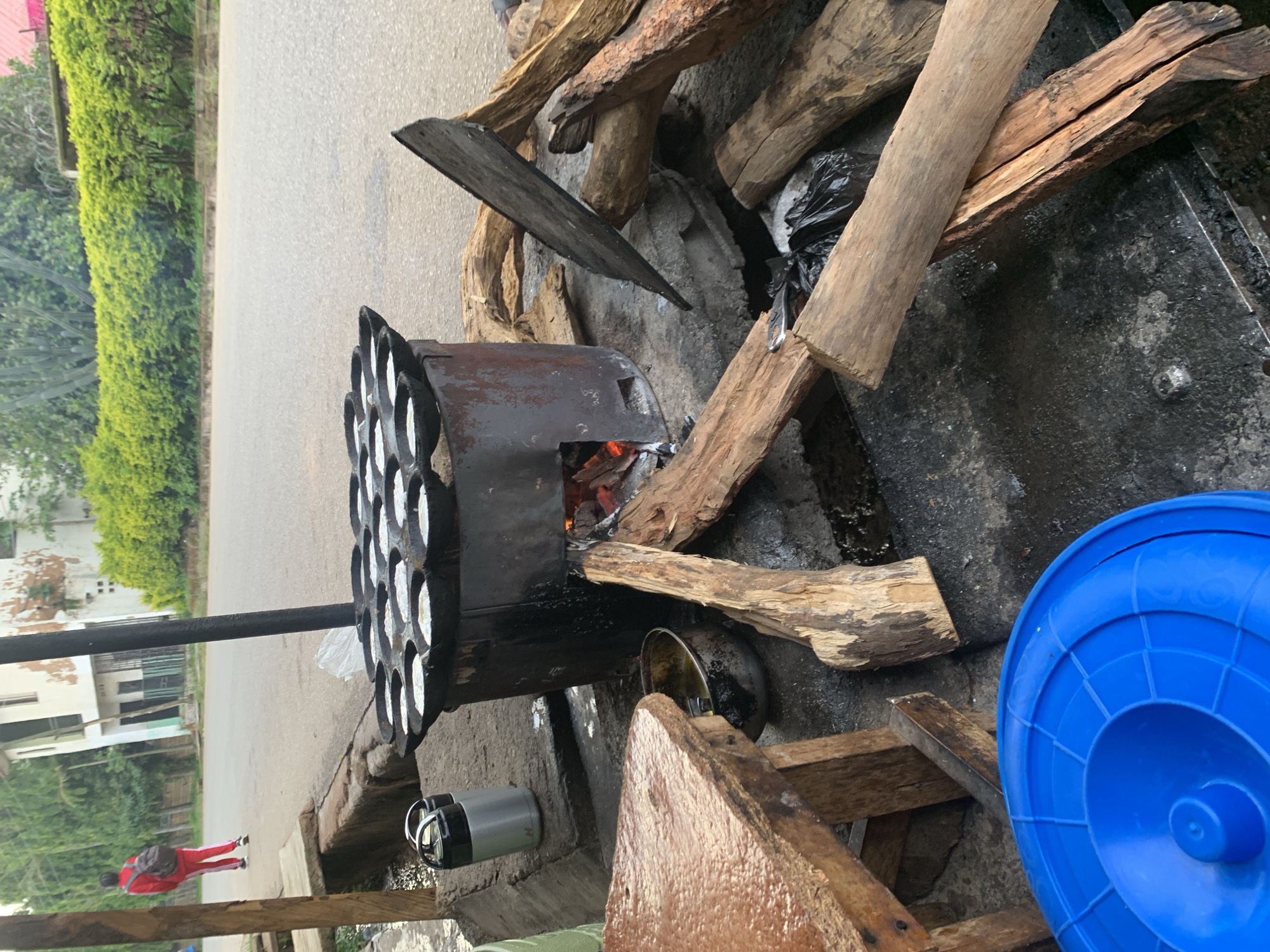 This image depicts a street food stall in Jos, Nigeria, on a cloudy day. In the foreground, a blue plastic lid rests on the ground, partially obscuring a weathered wooden table. Behind the table, a rustic iron stove is in operation, with visible flames fueling a cooking surface dotted with round molds containing a white batter, presumably for a local delicacy. Next to the stove, a metal pot sits with what appears to be cooking oil. A collection of chopped firewood is piled around the stove, ready for use. In the mid-ground, two insulated thermoses are positioned on the ground. Further back, a person wearing a red tracksuit and a backpack walks away from the camera on a paved road. The background features a building with white walls and green hedges lining the roadside, suggesting an urban environment. The overall scene captures a moment of everyday life and commerce on the streets of Jos.