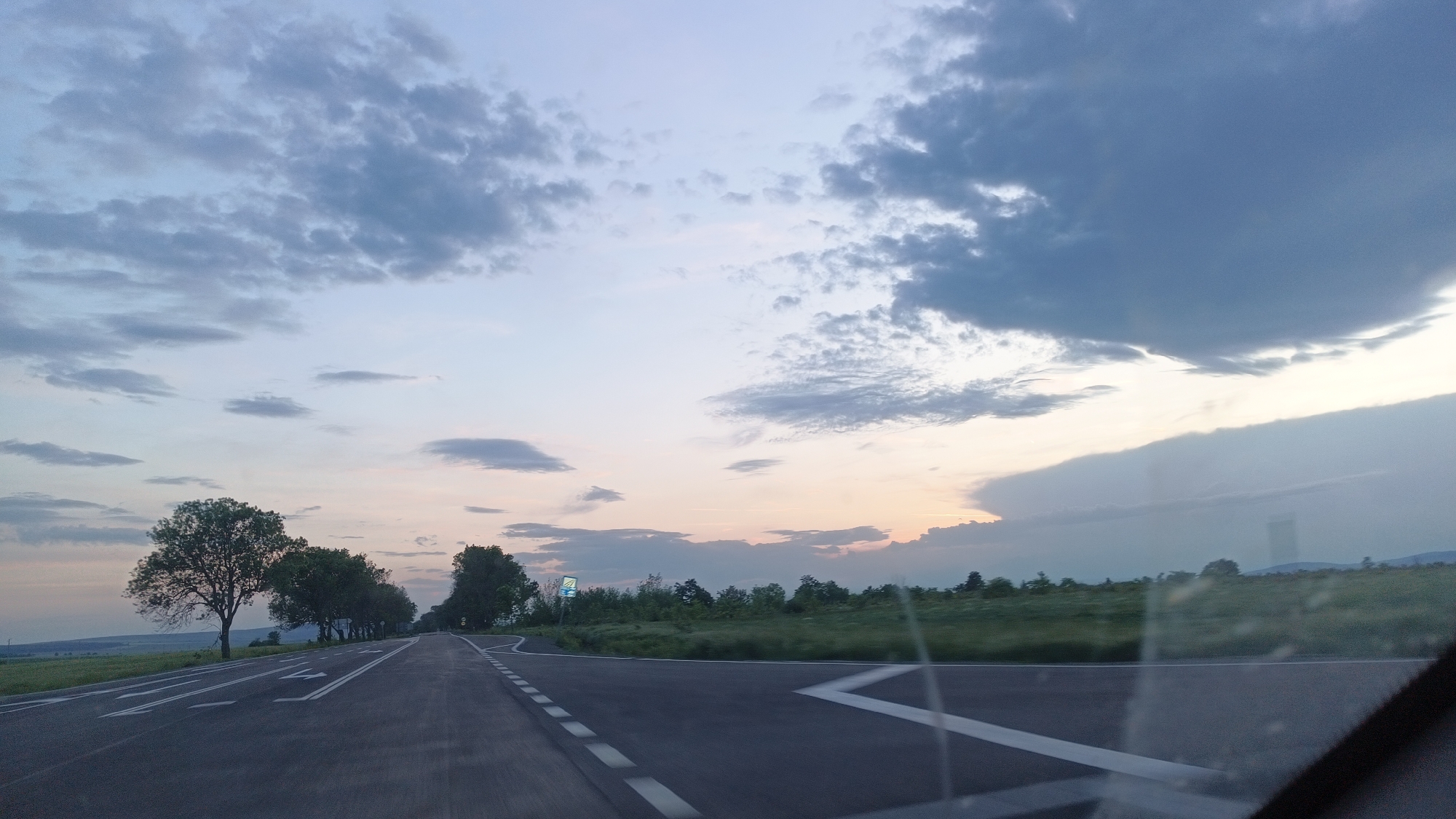 The image depicts a two-lane asphalt road in a rural area, likely near Sokolovo, Bulgaria. The road has white painted lane markings and is oriented toward the horizon. Trees line the left side of the road, casting long shadows. To the right of the road, a grassy field extends towards the horizon. The sky is filled with clouds, ranging from light gray to dark blue, suggesting either early morning or late evening light. The photograph appears to have been taken from inside a vehicle, indicated by the presence of the windshield's edge in the lower right corner. There are no people visible in the image.
