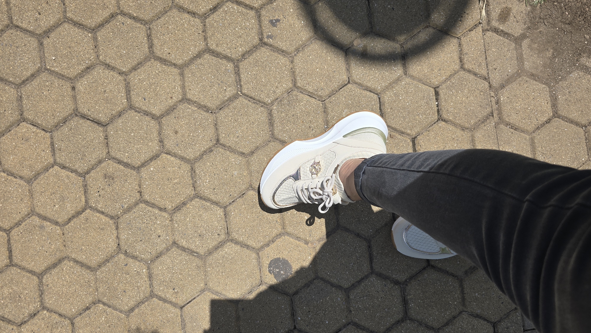 A first-person perspective shot shows a person standing on hexagonal concrete pavers. The person is wearing dark gray jeans and a light-colored sneaker with white laces and a tan sole. A shadow of the person is cast on the ground to the left. The setting appears to be an outdoor urban environment in Vratsa, Bulgaria, possibly a park or walkway. The lighting suggests a sunny day.
