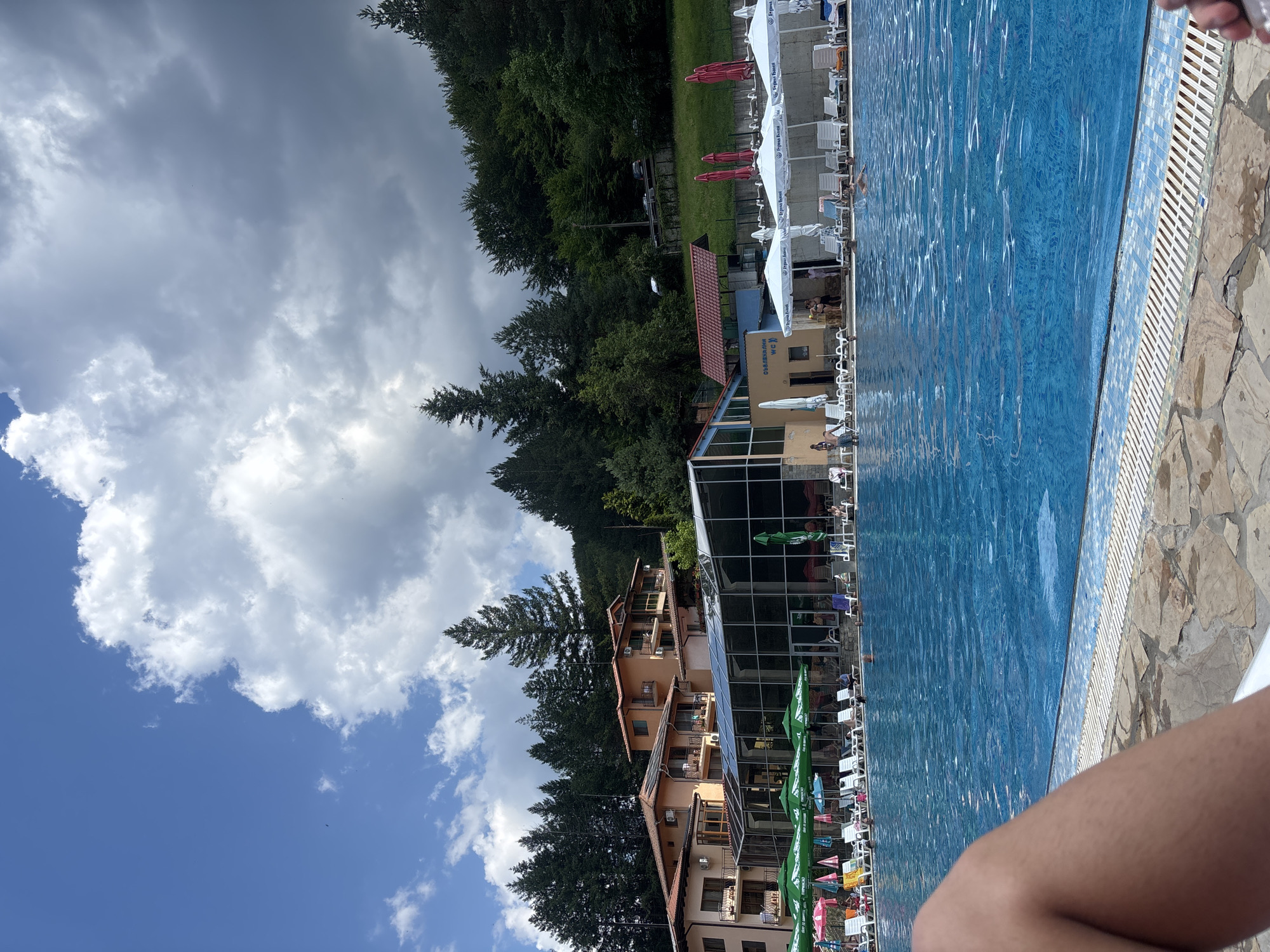 The image depicts an outdoor swimming pool area at a resort in Chiflik, Bulgaria. The foreground features the edge of the pool with stone and tile bordering, and a person's leg is visible at the lower left corner, suggesting the photo is taken from a lounging position. The pool itself is filled with clear blue water, with several people swimming or relaxing within it. Surrounding the pool, numerous white lounge chairs and umbrellas are set up, some green and pink umbrellas are also visible, indicating a recreational setting.
In the background, a modern building with a glass facade and a tiled roof is situated, likely part of the resort facilities. Beyond the building, a dense forest covers a hillside, with tall trees reaching towards a partly cloudy sky. The clouds are a mix of bright white and darker gray, suggesting variable weather conditions. The resort appears to be nestled in a natural, hilly environment, characteristic of the Chiflik region known for its thermal springs and mountain landscapes.