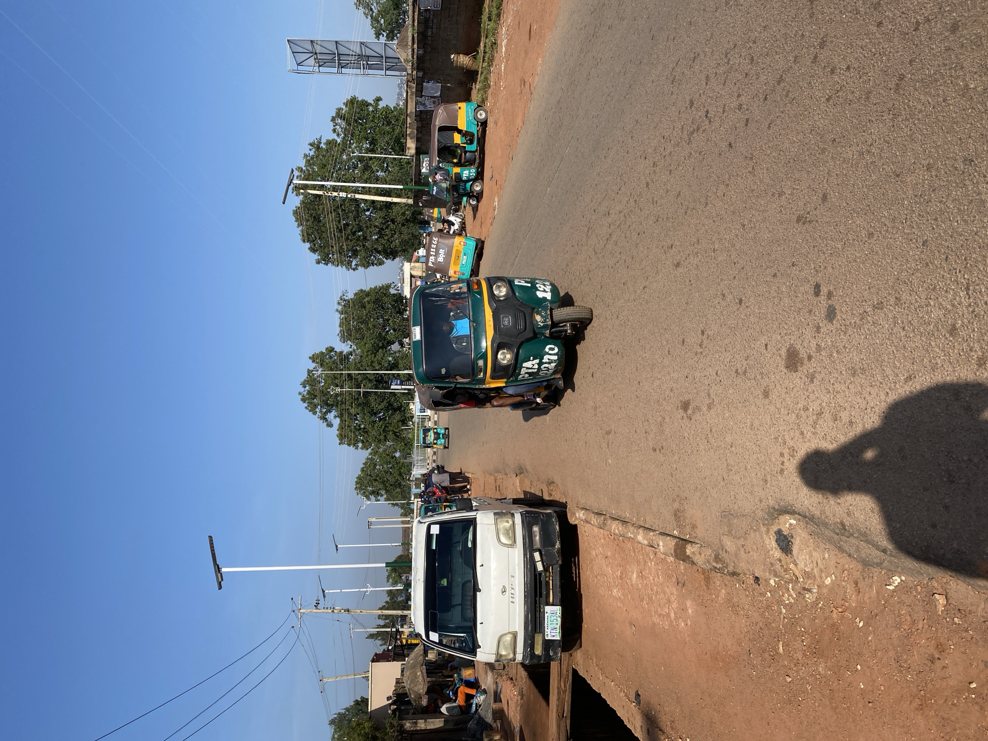 The image depicts a street scene in Bar Jirgi Summa, Nigeria, on a clear, sunny day. The road is paved with asphalt and bordered by a red dirt shoulder. Several vehicles are visible, including a white Hiace mini-truck parked on the side, a green and yellow auto-rickshaw in the foreground with "PTA-1270" visible on its side, and two other auto-rickshaws further down the road, one yellow and green with "PTA-11266 Bolt" on it, and another smaller green and yellow one. In the background, trees line the side of the road, and power lines and streetlights are present. A few people are visible walking and standing near the mini-truck and on the side of the road. The overall impression is of a typical bustling street in a Nigerian town.