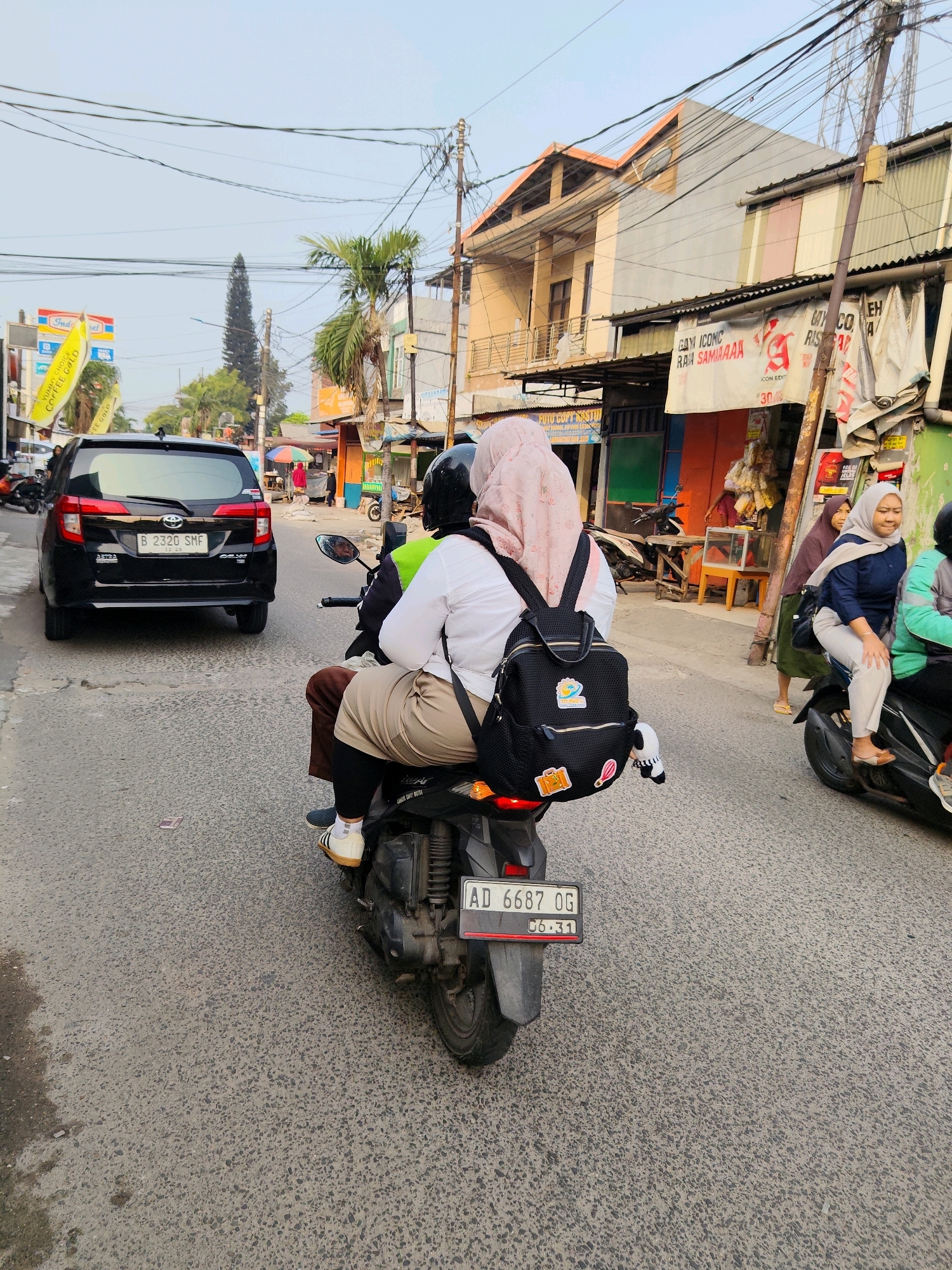 In a street scene in Bekasi, Indonesia, two people are riding on a motorcycle. The person in the back is wearing a pink hijab and a white shirt, carrying a black backpack with several patches. The person in the front is wearing a black helmet and a green jacket. They are riding on a paved road with a black car in front of them. Buildings line the street, with shops and signs visible. Overhead, a tangle of electrical wires crisscrosses the sky. The overall impression is a bustling urban environment.