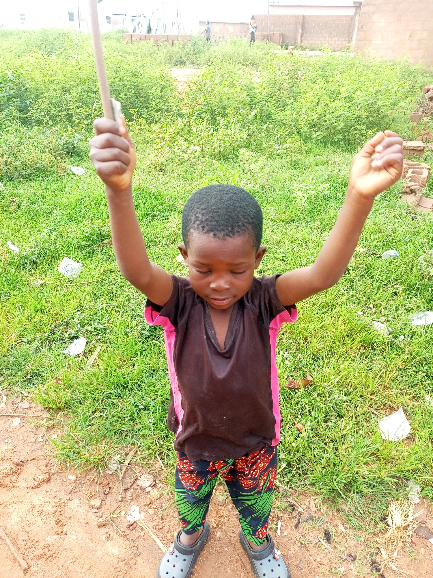 In Tempure, Nigeria, a young child with dark skin stands in a dirt and grassy area, holding a stick in their right hand above their head. The child is wearing a dark brown shirt with pink accents on the sleeves and sides, and patterned pants with red and green designs. The child's eyes are closed and their head is tilted down. Their arms are raised. The background consists of dense green vegetation, a brick wall, and some buildings. The ground is a mix of reddish-brown dirt and green grass. Scattered pieces of white debris are visible on the grass.