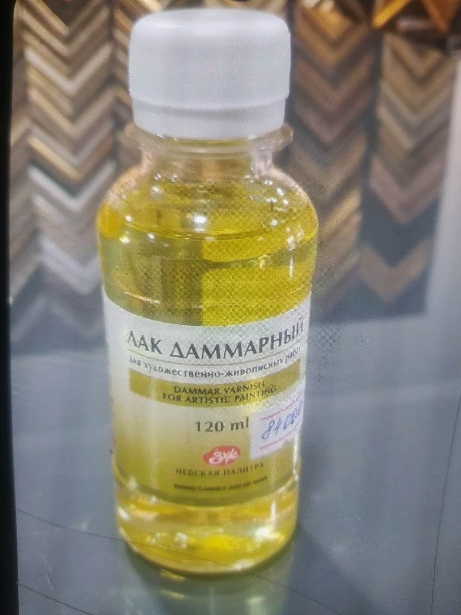 The image is a close-up, slightly angled, eye-level shot of a bottle of Dammar varnish. The bottle is clear plastic and filled with a yellowish liquid. A white screw-on cap seals the bottle. A label wraps around the lower portion of the bottle. The label is primarily white with text in Russian and English. The Russian text on the label appears to read "ЛАК ДАММАРНЫЙ" and "для художественно-живописных работ". The English text reads "DAMMAR VARNISH FOR ARTISTIC PAINTING". Below this, the volume is indicated as "120 ml". A stylized logo, possibly an artist's palette or a similar symbol, is visible in red. The price "84000" is handwritten in blue marker on the label. The background is blurred but appears to be a display of framed artwork with a chevron pattern of brown and gold tones. The scene is indoors, likely in an art supply store or gallery. There are no people visible in the image. The bottle rests on a gray surface, possibly a countertop or display shelf. The overall lighting is even and appears to be from artificial sources. There is no discernible indication of the specific location within the city of Birlik, Uzbekistan.