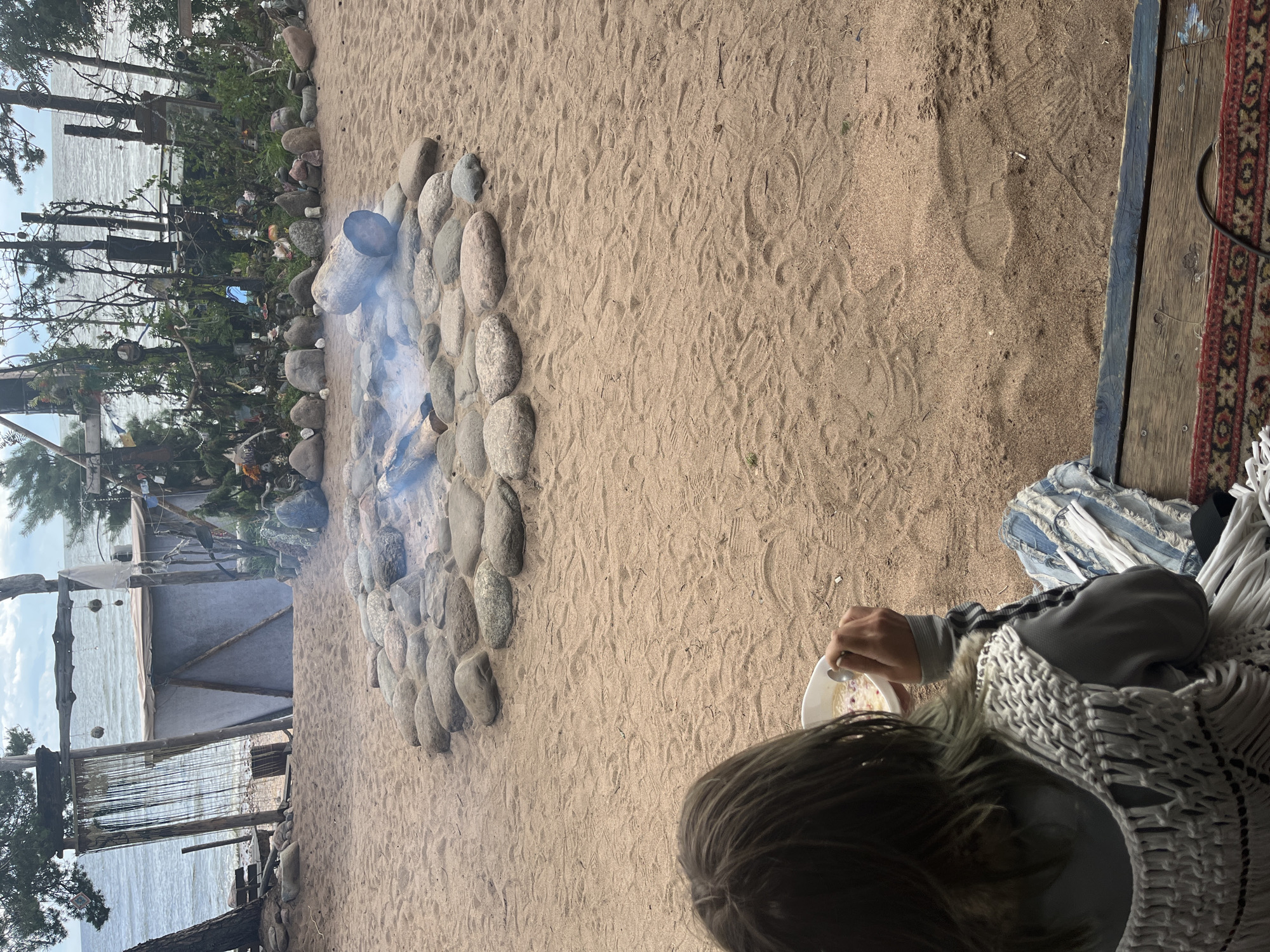 A person sits on a wooden deck facing a sandy area with a fire pit. The fire pit is circular and made of stones, with burning logs in the center. Behind the fire pit, there are trees and a body of water. The person is holding a bowl and a spoon, appearing to eat. The environment is outdoors, with sand, rocks, and vegetation visible. The person is wearing a light-colored, fringed vest over a grey long-sleeved shirt and ripped denim pants.