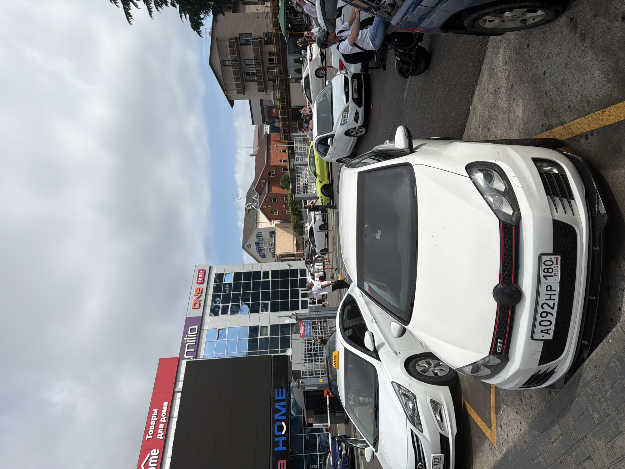 This is a medium shot of a busy street scene in Sochi, Russia, likely near a commercial establishment. A white Volkswagen Golf GTI, with license plate A092HP 180, is parked in the foreground, facing towards the camera and slightly to the right. The car has a black grille with red accents and a black emblem. Behind it, a white Hyundai sedan is partially visible on the left, with a taxi sign on its roof. Further back, several other cars are present, including a yellow sports car and a white sedan. A man in a white t-shirt and dark shorts is walking in the middle ground, carrying a yellow bag. People are visible in the background, some standing and others in vehicles. Buildings with retail signage, including "HOME" and "DNS ГИПЕР", are visible in the background. The sky is cloudy. The scene appears to be an outdoor parking lot or street in a commercial area.