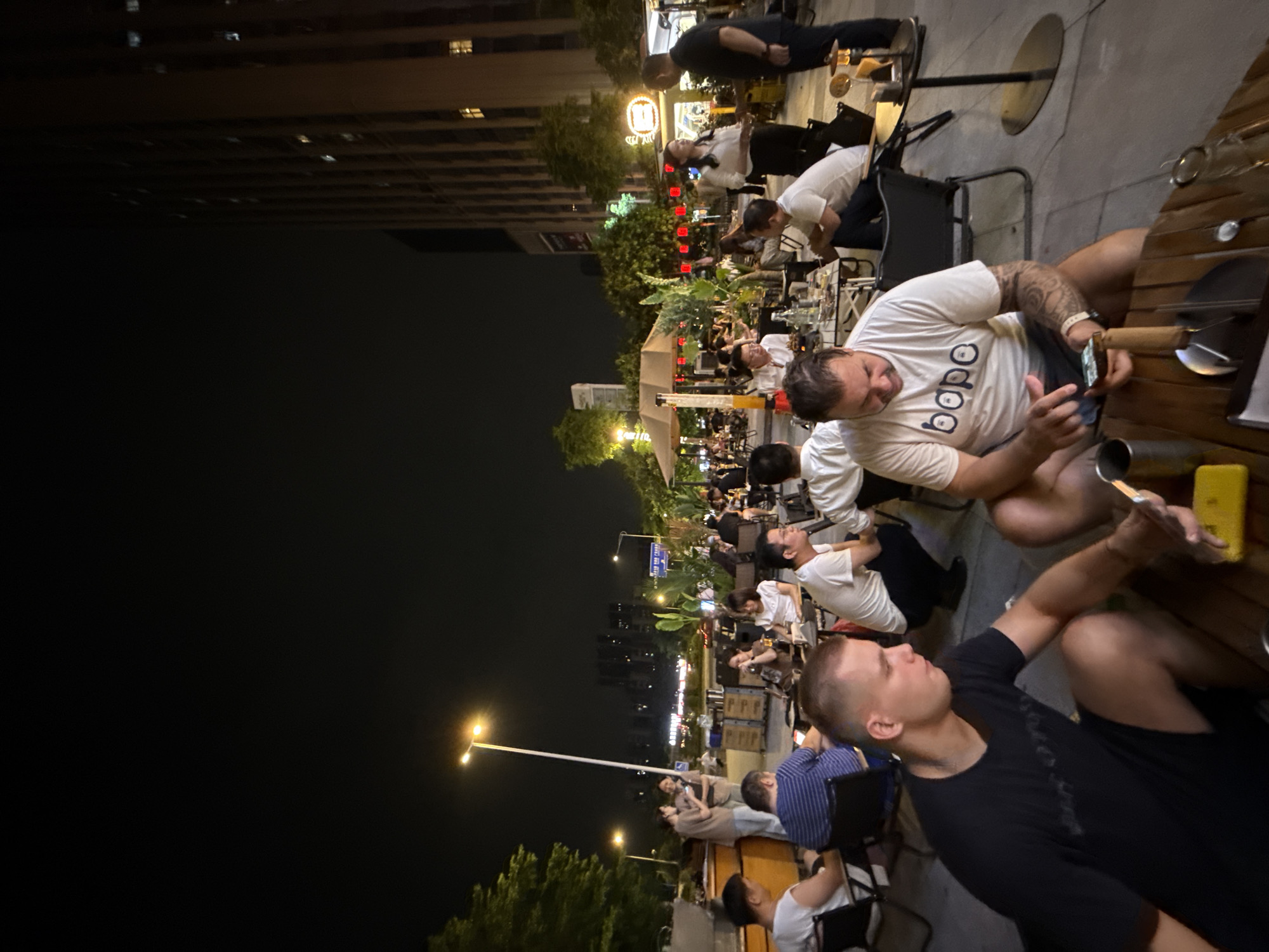 The image depicts a nighttime outdoor gathering of people at a restaurant or bar in Haizhu District, China. The scene is illuminated by streetlights and ambient lighting from the establishment. In the foreground, two men are seated at wooden tables. The man on the left wears a black t-shirt and shorts, while the man on the right wears a white t-shirt with "bobo" printed on it and shorts, and he is actively using a mobile phone. Numerous other individuals are seated at tables and standing around the outdoor seating area, some engaged in conversation and others appearing to be dining or drinking. In the background, a tall building, trees, and other urban elements are visible. The overall atmosphere suggests a social and casual evening setting.