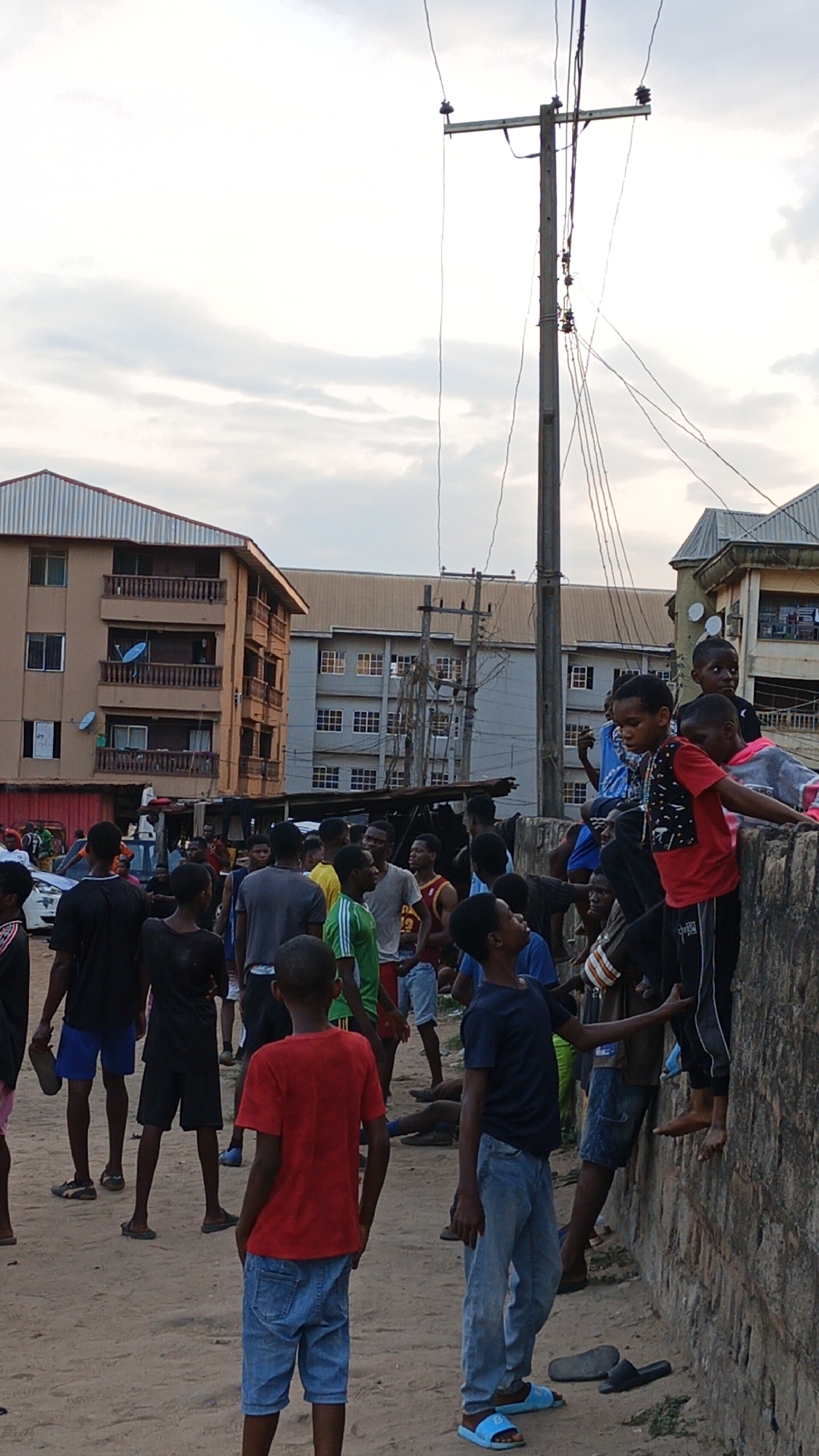 This image depicts a vibrant street scene in Onitsha, Nigeria, primarily featuring a large group of young males, ranging from children to teenagers. The setting is an outdoor, urban or semi-urban environment with a prominent dirt road.
In the foreground and midground, numerous individuals are standing, sitting, and interacting informally. Many are dressed in casual attire, including t-shirts, shorts, and jeans. On the right side, several children are perched on a low stone or concrete wall, observing the surroundings. The overall mood appears casual and social.
The background is filled with multi-story buildings characteristic of a residential area. A brown apartment building with multiple balconies and satellite dishes is visible on the left, while a larger, grey building stands further back. A tall, weathered wooden utility pole with numerous electrical wires and insulators dominates the right side of the frame, indicating the local infrastructure.
The time of day is likely late afternoon or early evening, evidenced by the soft, diffused light and the partly cloudy or overcast sky. This light quality suggests the sun is not high in the sky, creating a gentle illumination over the scene. There is no discernible text visible on any of the subjects or objects within the image.