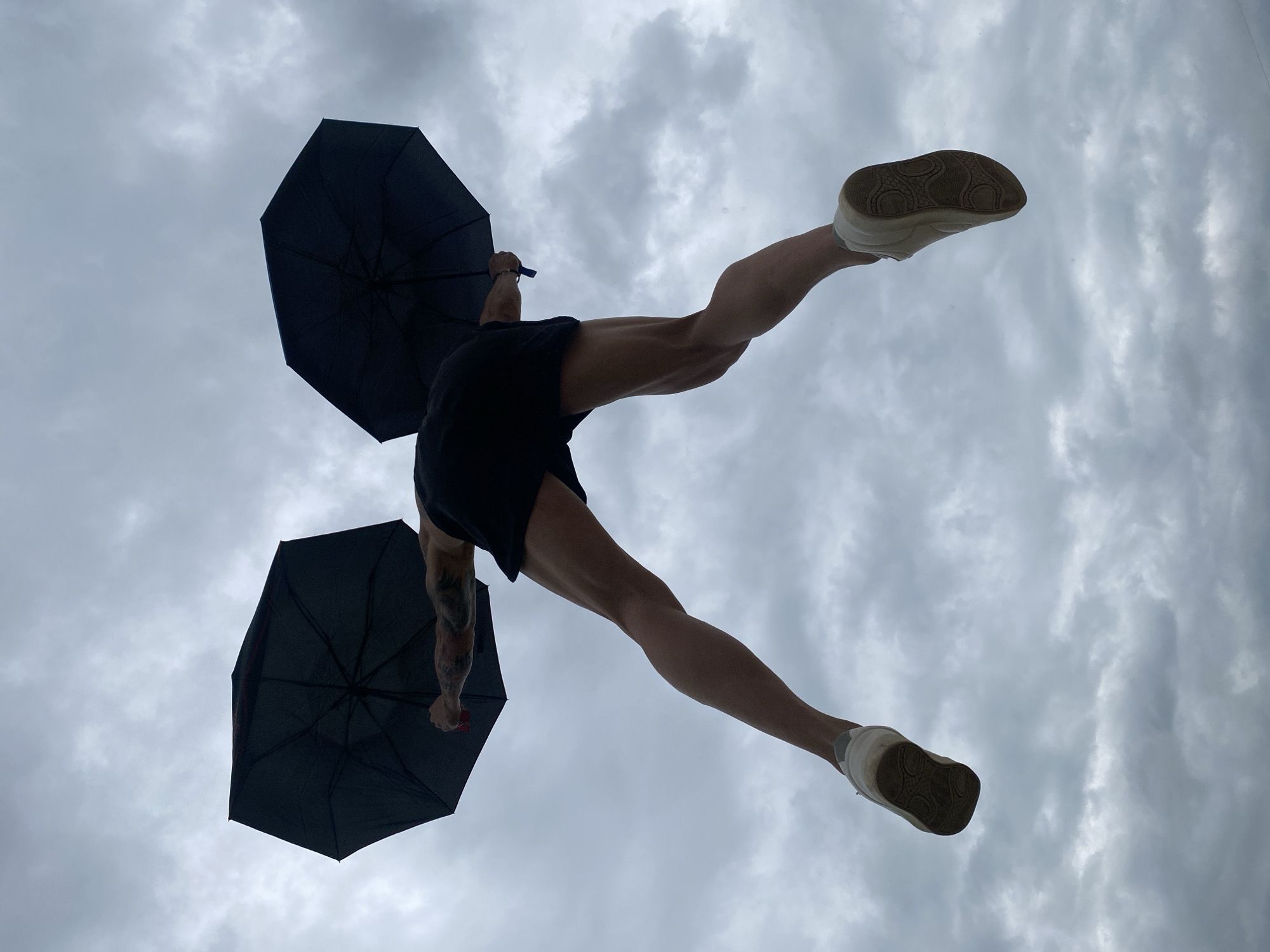An individual is captured mid-air against a cloudy sky, holding two open black umbrellas, one in each hand. The person is wearing black shorts and white sneakers. The perspective is from below, looking up, which emphasizes the person's muscular legs and the position of the umbrellas. The background consists of an overcast sky with diffuse cloud cover. The location is the city of Pleven, Bulgaria.