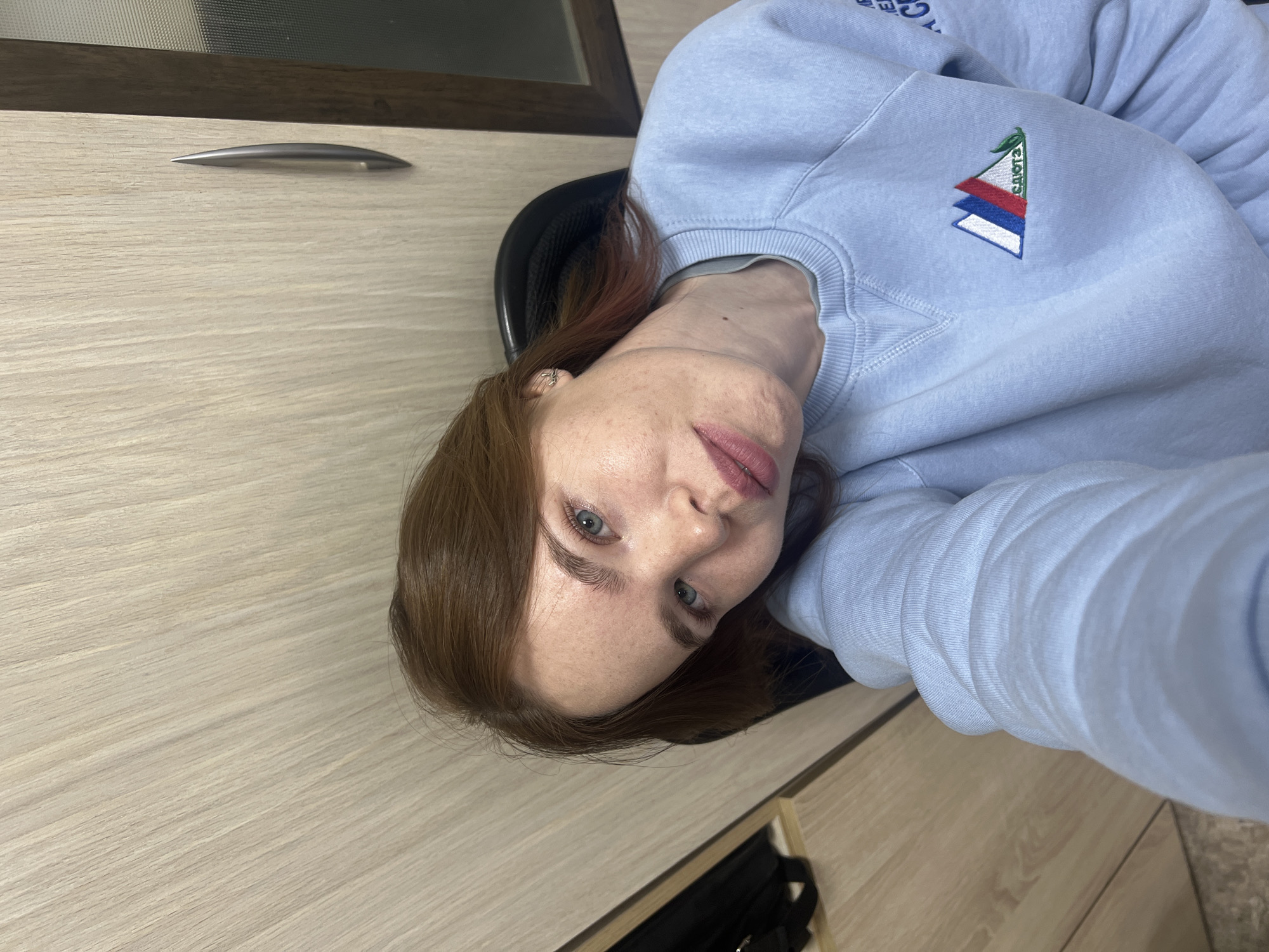 A person with light brown hair and blue eyes is sitting in a black chair facing the camera. They are wearing a light blue sweatshirt with a triangular patch featuring red, white, and blue stripes along with the word "Salut" embroidered on it. The person is indoors, possibly in an office setting, as indicated by a light wooden desk with a silver handle. The location is presumed to be Chaikovsky, Russia, based on the "Salut" patch which may be associated with that city.