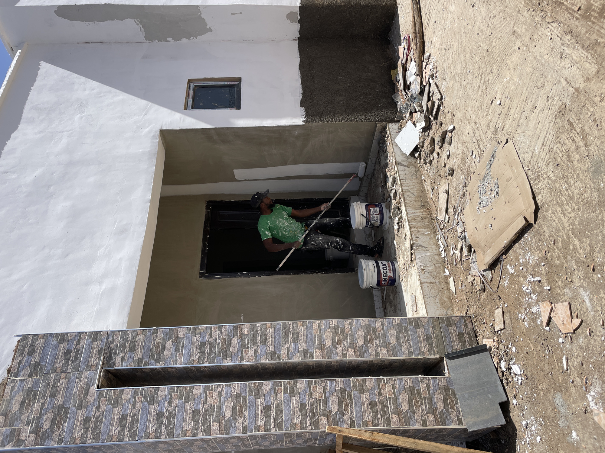 A construction worker is painting the ceiling of an outdoor entryway, located outside a house in Jos, Nigeria. The building's exterior is a mix of white paint and grey stone cladding. The entryway ceiling is painted white, and the worker, wearing a green t-shirt, black trousers covered in white paint spots, and a black cap, is using a paint roller attached to a long pole to apply a fresh coat. Two paint buckets labeled "FINECOAT Acrylic Emulsion" are positioned near him on the ground. The ground around the building is dirt with construction debris including tile pieces and a cardboard panel.