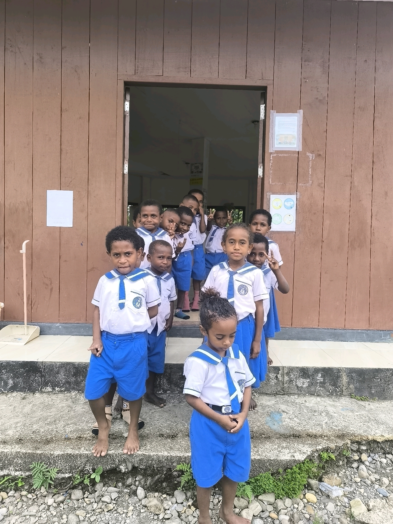 A group of young students, all wearing matching blue shorts and white shirts with blue trim and ties, stand in front of a wooden building. The building appears to be a school, with a doorway in the center leading into a dimly lit interior. Several of the students are looking directly at the camera, while others are looking off to the side. One student in the foreground is looking down, with their hands clasped. The students are standing on a concrete step that leads down to a rocky ground. There is a broom and dustpan leaning against the side of the building to the left of the doorway. On the exterior wall of the building, to the right of the doorway, there are two pieces of paper taped up. The overall scene suggests a school environment in a tropical or subtropical location.