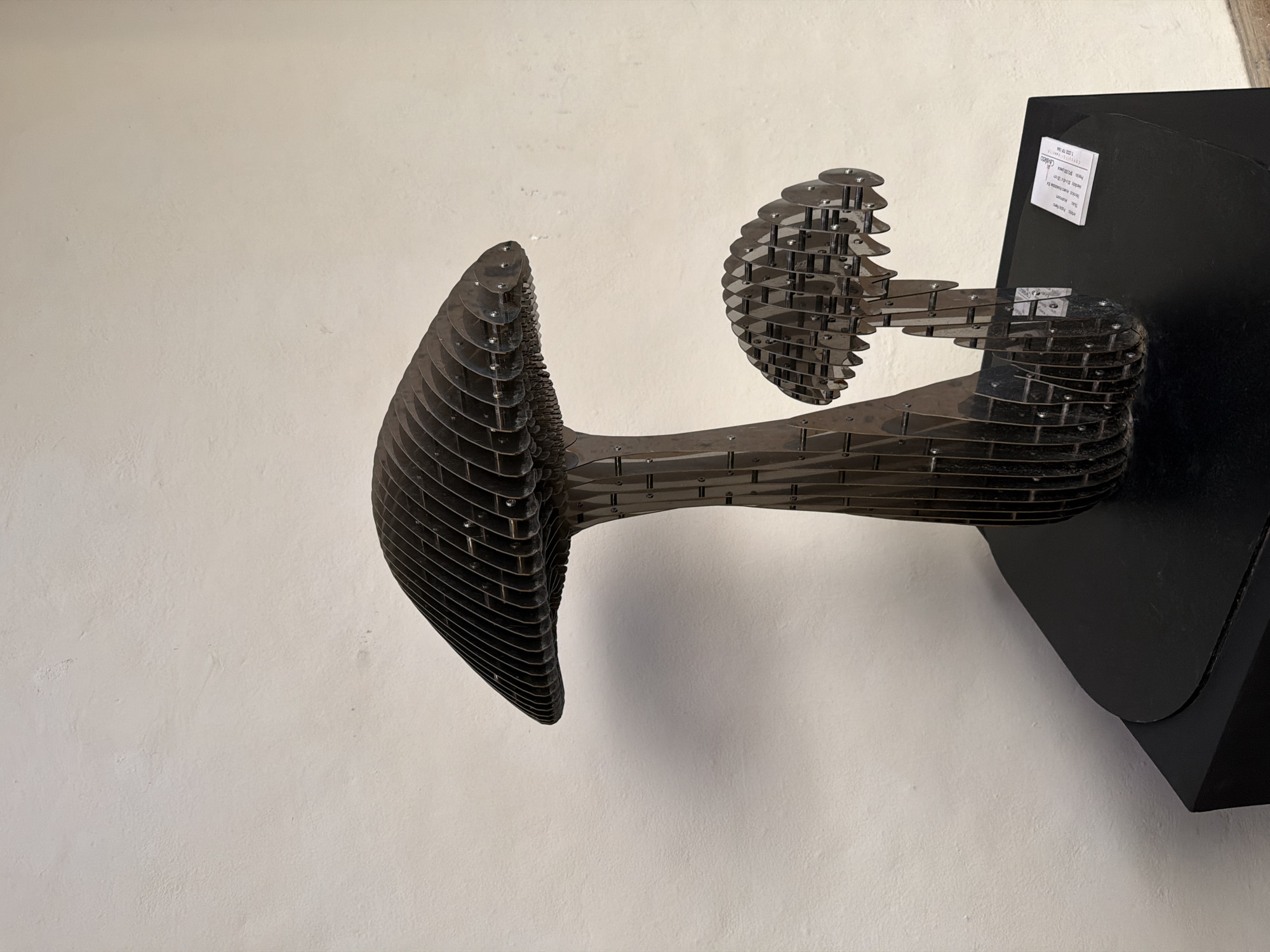 A close-up shot presents an abstract metal sculpture mounted on a black base against a plain white wall. The sculpture consists of multiple thin, horizontal metal plates stacked to form two bulbous shapes connected by a cylindrical body. The shape on the left is larger and more elongated, resembling a stylized head, while the one on the right is smaller and rounder. A white label with text is affixed to the black base. No people are visible. The location, San Bernardino Tlaxcalancingo, Mexico, cannot be ascertained from this photograph alone.