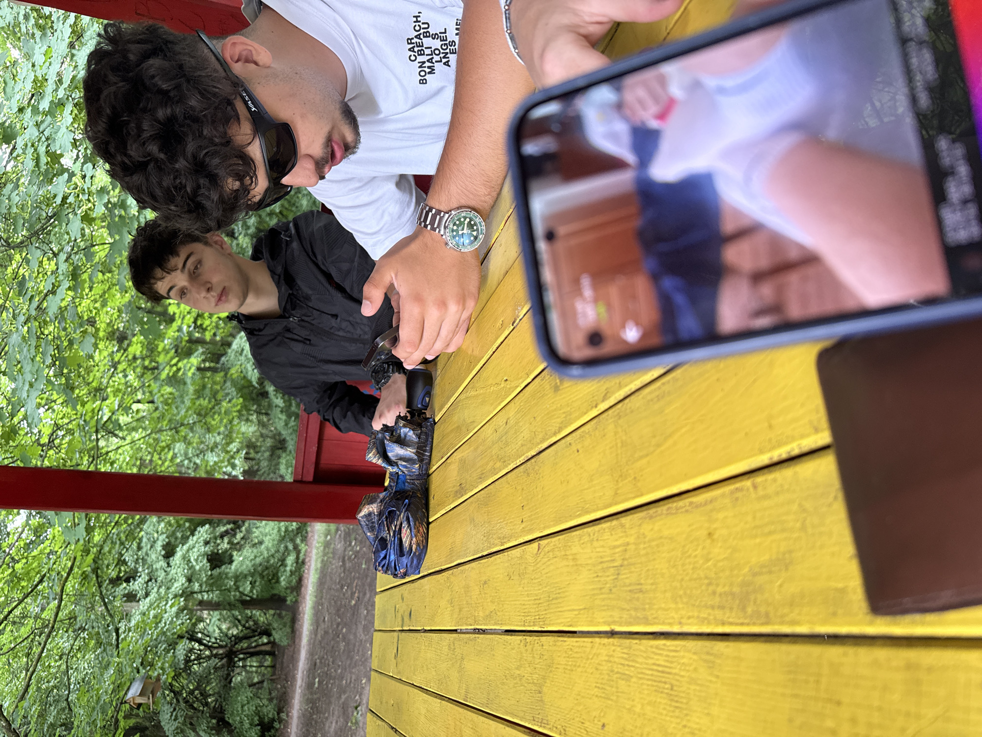 The image captures a casual outdoor scene in what appears to be a park or wooded area, with Sofia, Bulgaria as the location context. Three young men are visible, seated or standing around a bright yellow slatted wooden table or bench.
In the mid-ground, a man with curly dark hair and a beard wears black sunglasses and a white t-shirt. The text "BONEAR BEACH, MALIBU LOS ANGELES" is partially visible on his shirt. He is looking downwards, possibly at an object on the table. On his left wrist, he wears a prominent green-faced wristwatch with a metal band.
To his left, slightly further back, another man with short dark hair is looking upwards and to the right, towards the general direction of the first man's gaze. He is wearing a dark, possibly black, jacket. An umbrella or a patterned blue and black bag is seen on the table in front of him.
In the immediate foreground, partially visible on the right, is a hand holding a smartphone. The phone's screen, in sharp focus, displays a blurred image of a person, likely a female, from the waist down, wearing white clothing or socks against a wooden background. The person holding the phone is largely out of frame.
The setting includes lush green foliage and trees in the background, suggesting a natural environment. A red horizontal beam or support structure is visible behind the men. The lighting indicates it is daytime, with soft, diffused light, possibly due to an overcast sky or shade from trees. The overall mood appears relaxed and informal.