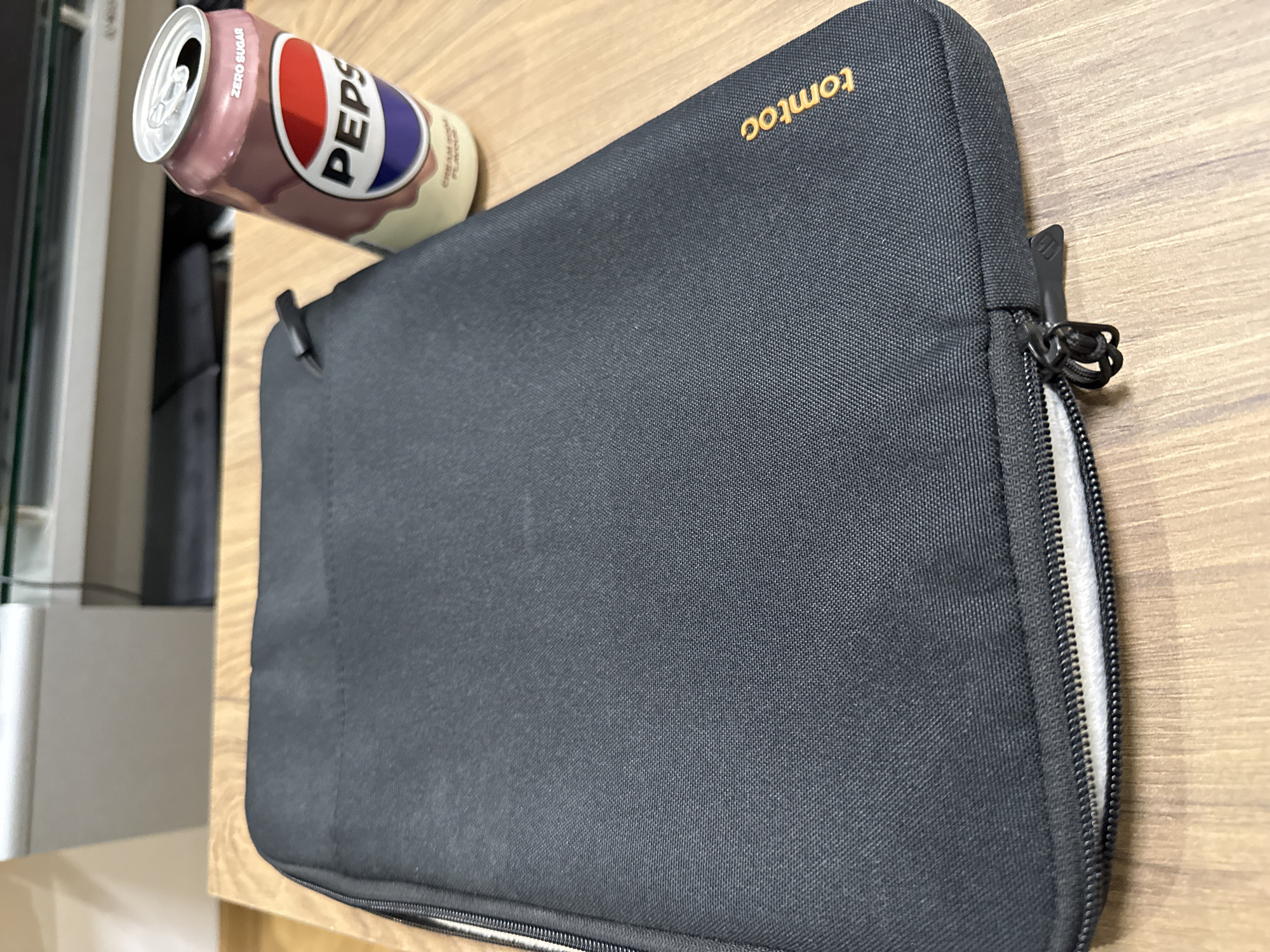 The image shows a wooden surface with a black laptop sleeve and a can of Pepsi Cream Soda. The laptop sleeve is partially unzipped, revealing a white interior. The "tomtoc" logo is visible on the sleeve. The can of Pepsi is partially obscured. The location is not specified in the image.