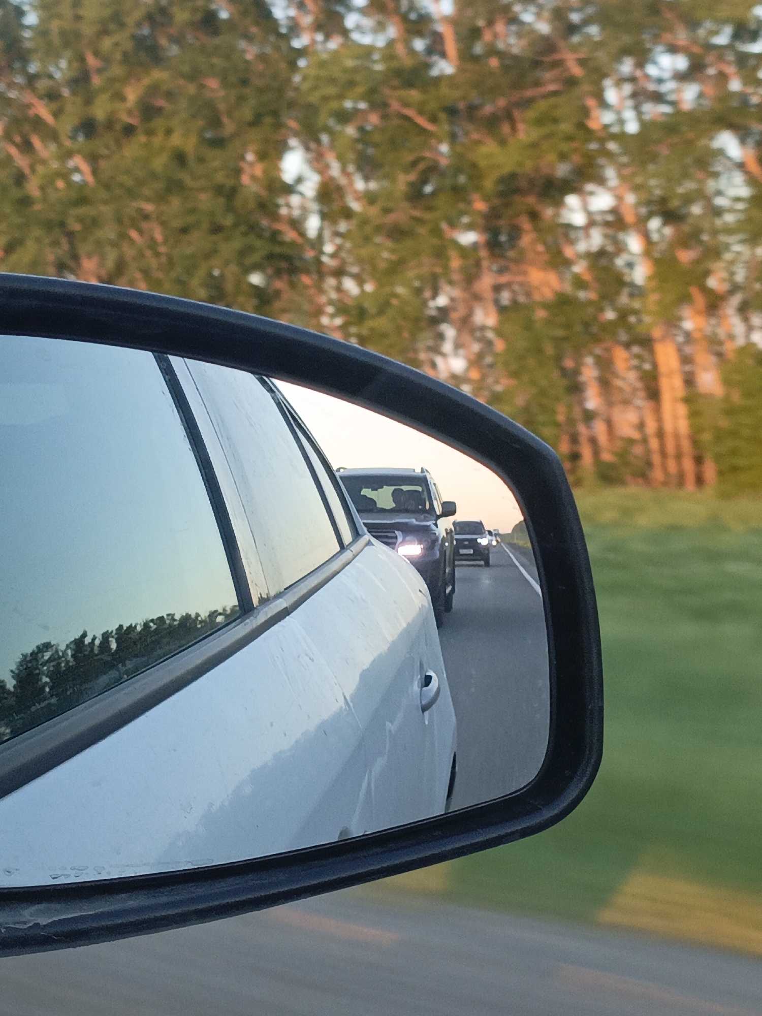 The image shows a side mirror of a white car. The mirror reflects the road behind the car, with several vehicles following. A forest is visible in the background. The location is likely in a rural area of the Ural region of Russia, as suggested by the presence of birch trees and the name of the city. The scene is from the perspective of the driver of the white car.
