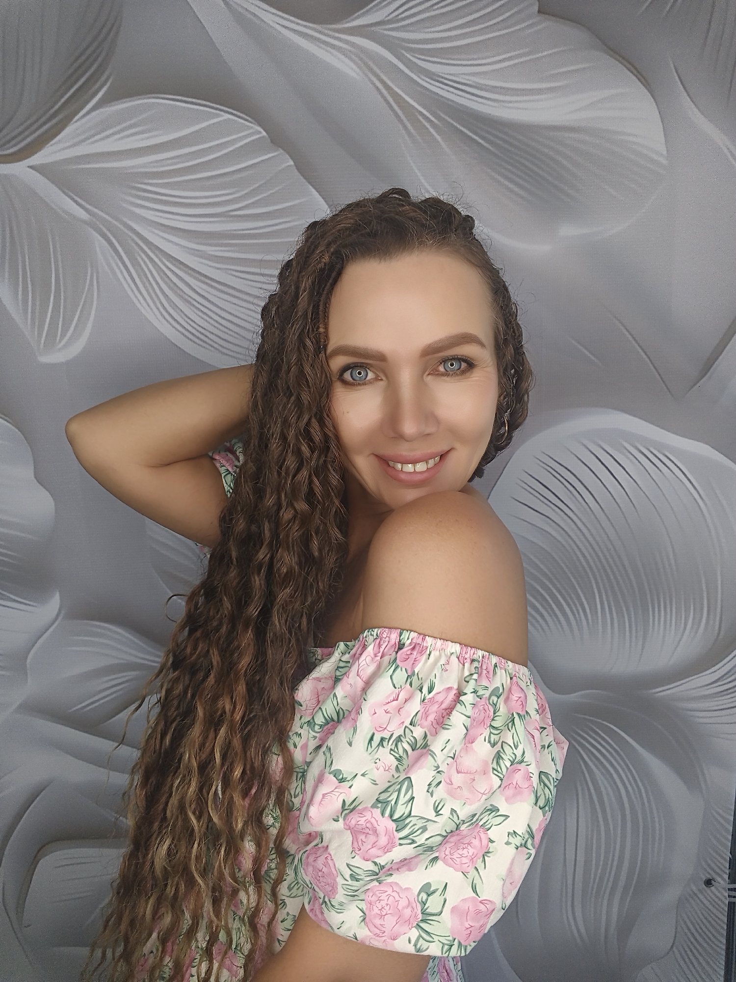 A woman with long, curly brown hair and bright blue eyes is shown from the chest up, leaning her head on her right arm. She is wearing an off-the-shoulder dress with a floral pattern of pink roses and green leaves against a white background. Her left arm is bent, with her hand behind her head. She is smiling, and her head is tilted slightly. The background is a textured, light gray wall with a subtle, abstract pattern of curved lines that resemble flower petals or feathers. The lighting is soft, illuminating the woman's face and upper body. The image appears to be a selfie taken indoors, with no specific city or location details discernible.