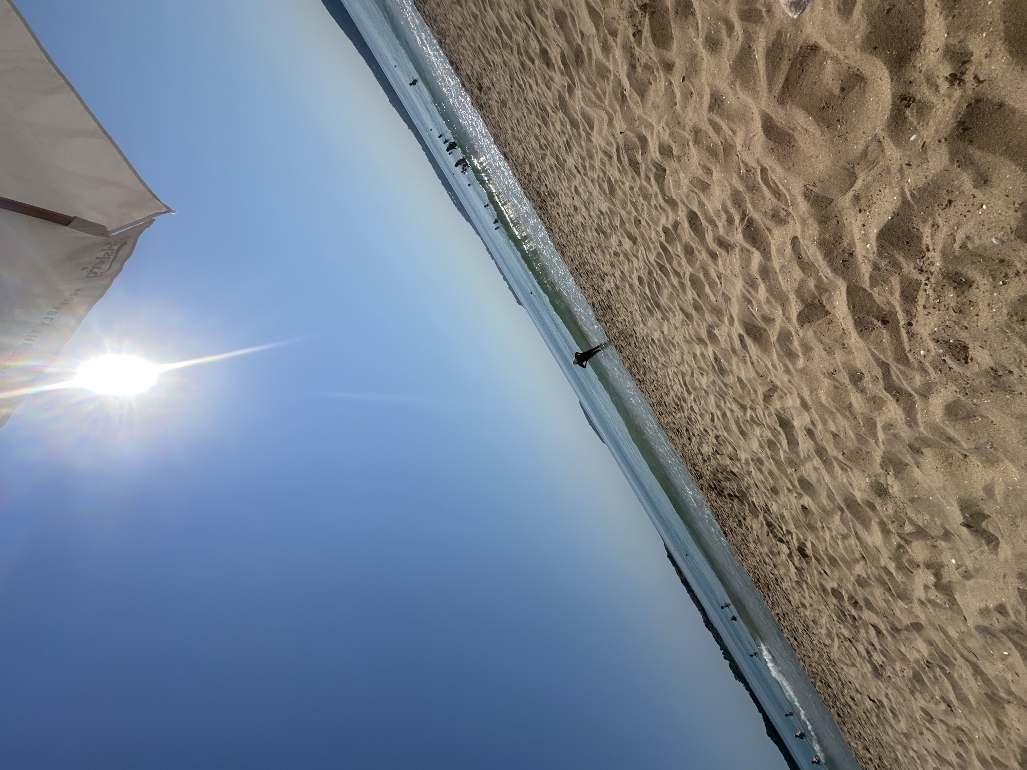 A beach scene in Sozopol, Bulgaria, features golden sand in the foreground with footprints and ripples. The clear blue sea stretches to the horizon, with a gentle wave breaking near the shore. Several people are visible in the water, some swimming and others wading. A single figure stands in the shallow water on the left, facing the sea. The sky is a bright, cloudless blue, with the sun prominently displayed near the top center, emitting bright rays. A portion of a white beach umbrella with visible text is present in the upper right corner of the image, casting a slight shadow. The overall impression is a sunny day at the coast.