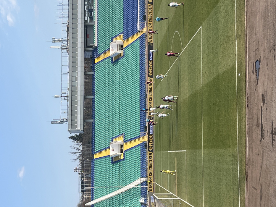 An outdoor football match is depicted at Vivacom Arena Georgi Asparuhov Stadium in Sofia, Bulgaria, under a clear blue sky. The meticulously striped green grass pitch occupies the midground, marked with white lines defining the penalty box, goal area, and a segment of the center circle. In the left foreground, a white goal frame is partially visible. Approximately 18 individuals are present on the pitch: a goalkeeper in a yellow kit positioned near the left goalpost, around 8 players in white jerseys with green accents, 8 players in maroon jerseys, and one referee in a black kit. Players are dispersed across the field's defensive and midfield sections, indicating active play. The background features empty stadium seating with distinct sections of green and blue bucket seats, separated by yellow access staircases. A horizontal advertising banner with "ОТВОРИ СИ СОБСТВЕН efbet ПУНКТ" and the efbet logo is displayed along the lower stand. Above the seating, a modern stadium structure with a grey facade and a roof equipped with several communication antennae is visible. The immediate foreground shows a grey concrete surface with dark, irregular stains.