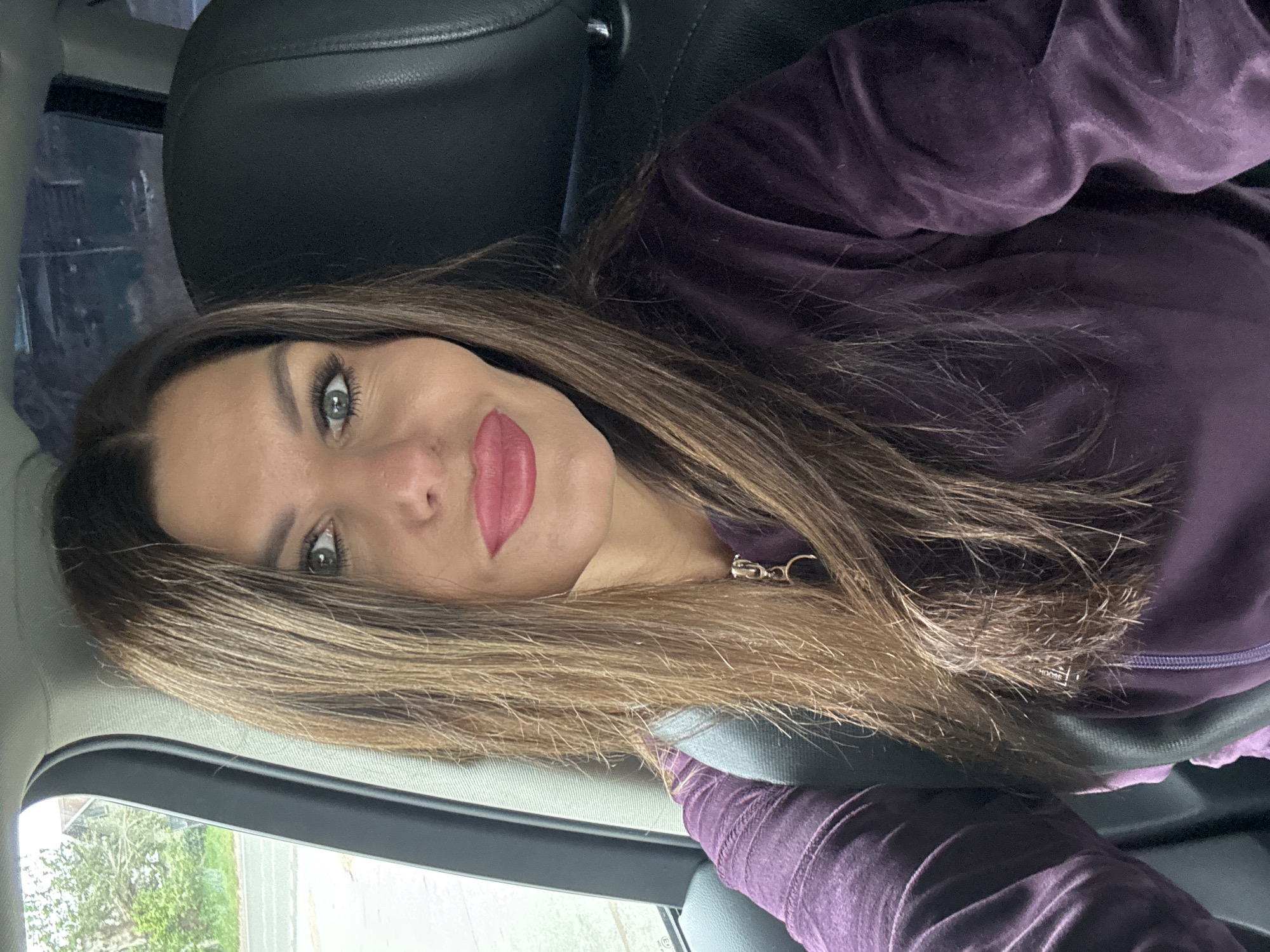 A woman with long, brown hair and full lips painted with pink lipstick is sitting in the driver's seat of a car. She is wearing a purple velour tracksuit and has a seatbelt fastened across her chest. Her gaze is directed towards the camera. The background shows a glimpse of a tree-lined street outside the car window, suggesting an urban or suburban setting. The interior of the car is dimly lit, with the focus on the woman's face and upper body.