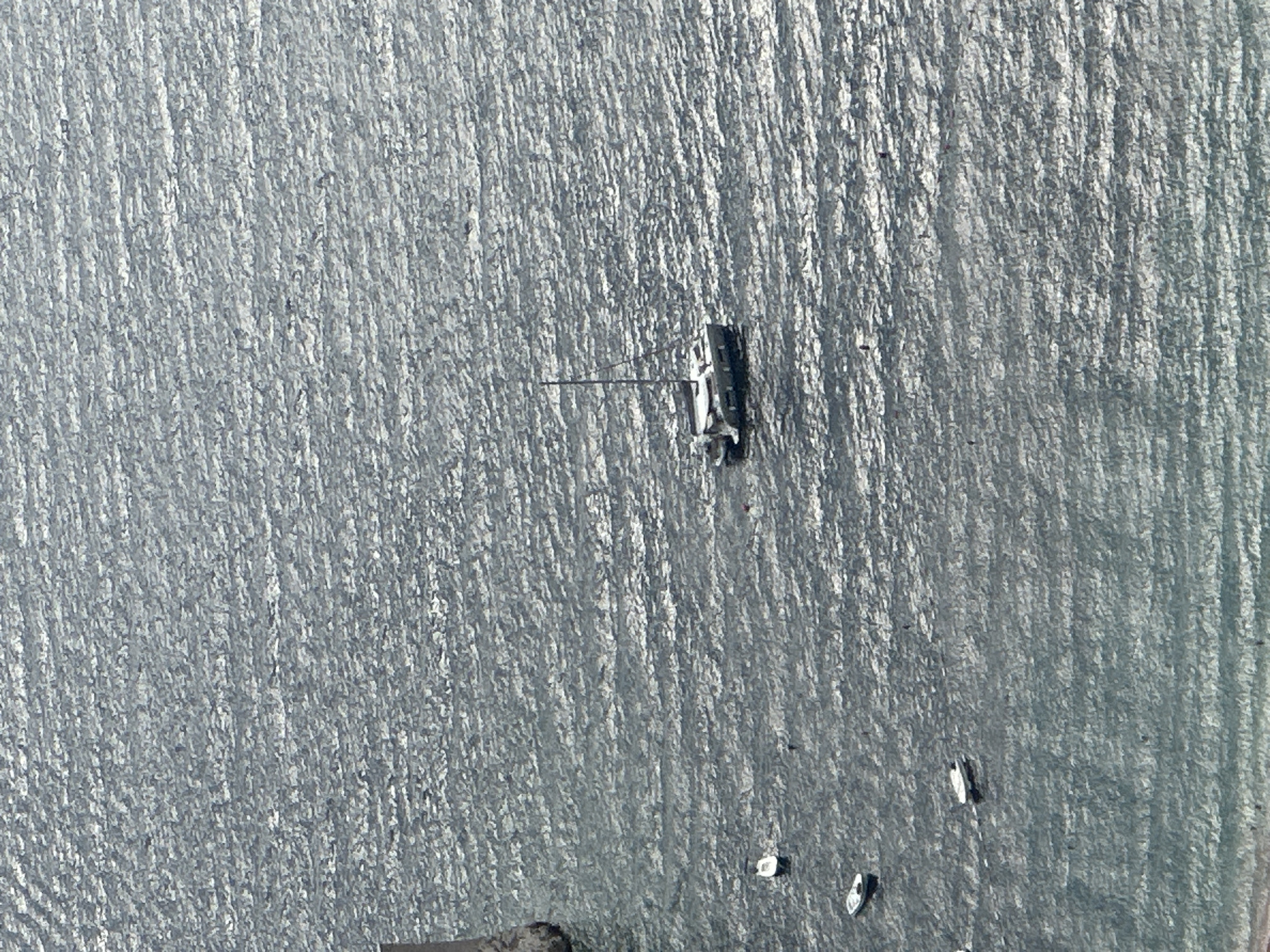 This is an aerial, high-angle drone photograph of the waters near Sveti Stefan, Montenegro. The sea is choppy with sunlight reflecting off the waves, creating a shimmering effect across the water. There are several small boats scattered on the water. In the center of the frame, a larger catamaran sailboat with two masts is anchored. To the left, a portion of the Sveti Stefan island and its causeway is visible at the edge of the frame. The lower portion of the image shows the shoreline with a sandy beach. The scene is devoid of human presence on the boats or the beach.