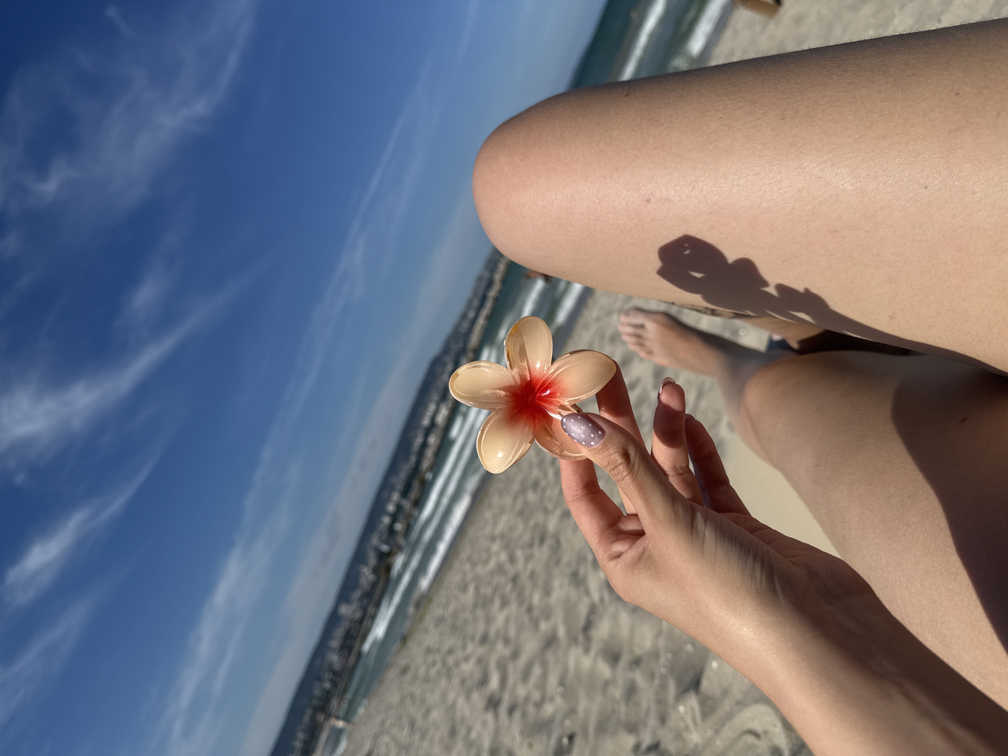 A woman's left hand holds a light pink and red flower with five petals, her fingernails painted with a light purple and white speckled design. Her bare feet are visible in the lower left quadrant of the image, with a shadow of the flower she is holding cast on her right thigh. She is sitting on a sandy beach with the ocean and a distant cityscape under a bright blue sky with wispy white clouds. The woman's legs are visible, with her right leg taking up a significant portion of the right side of the frame. The image is taken from a first-person perspective.