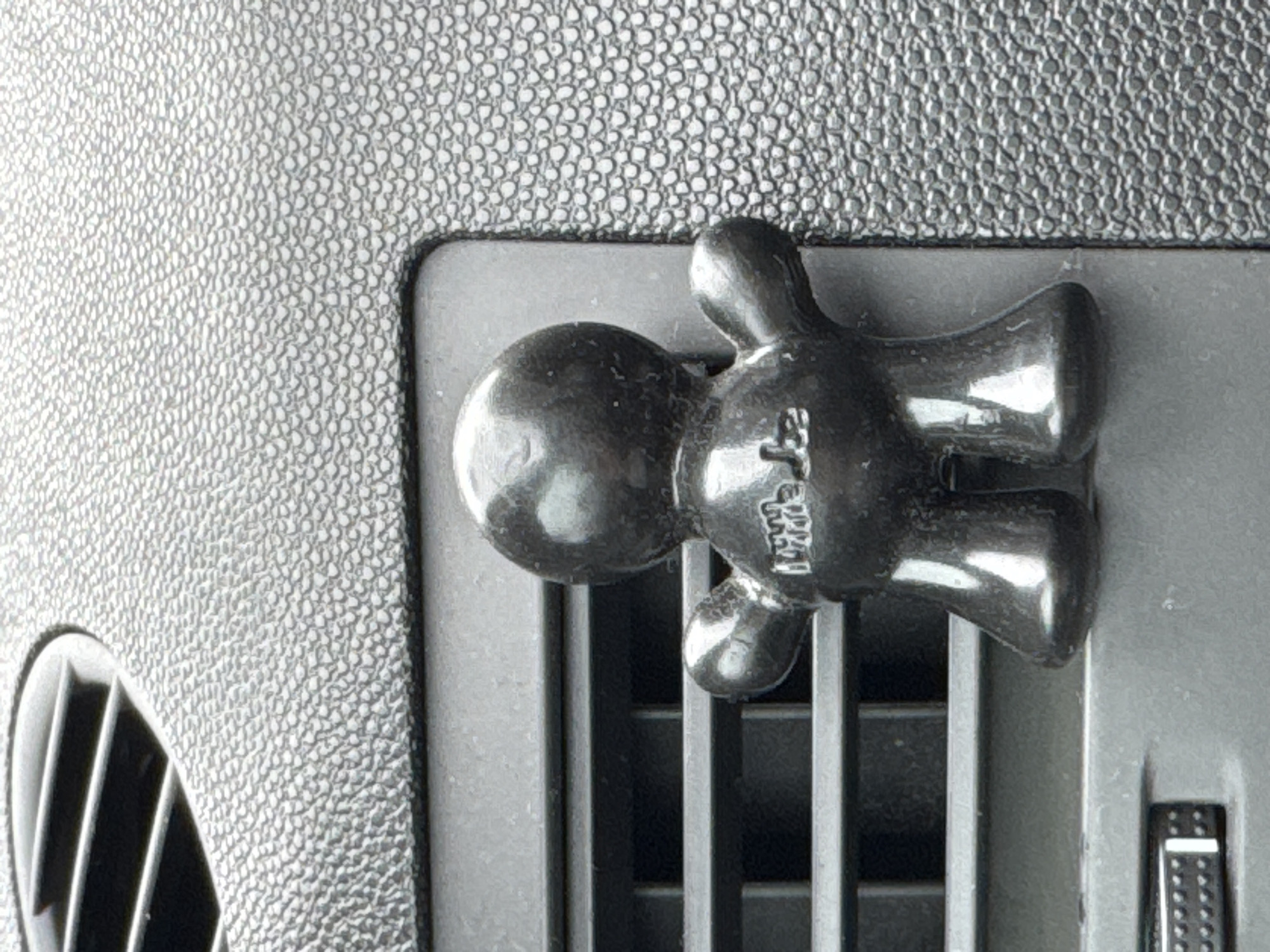 The image depicts a black, stylized human-shaped air freshener attached to a car's air vent. The air freshener appears to be made of plastic and has a glossy finish. The background shows a textured grey surface, likely the dashboard of the car. The location is unknown.