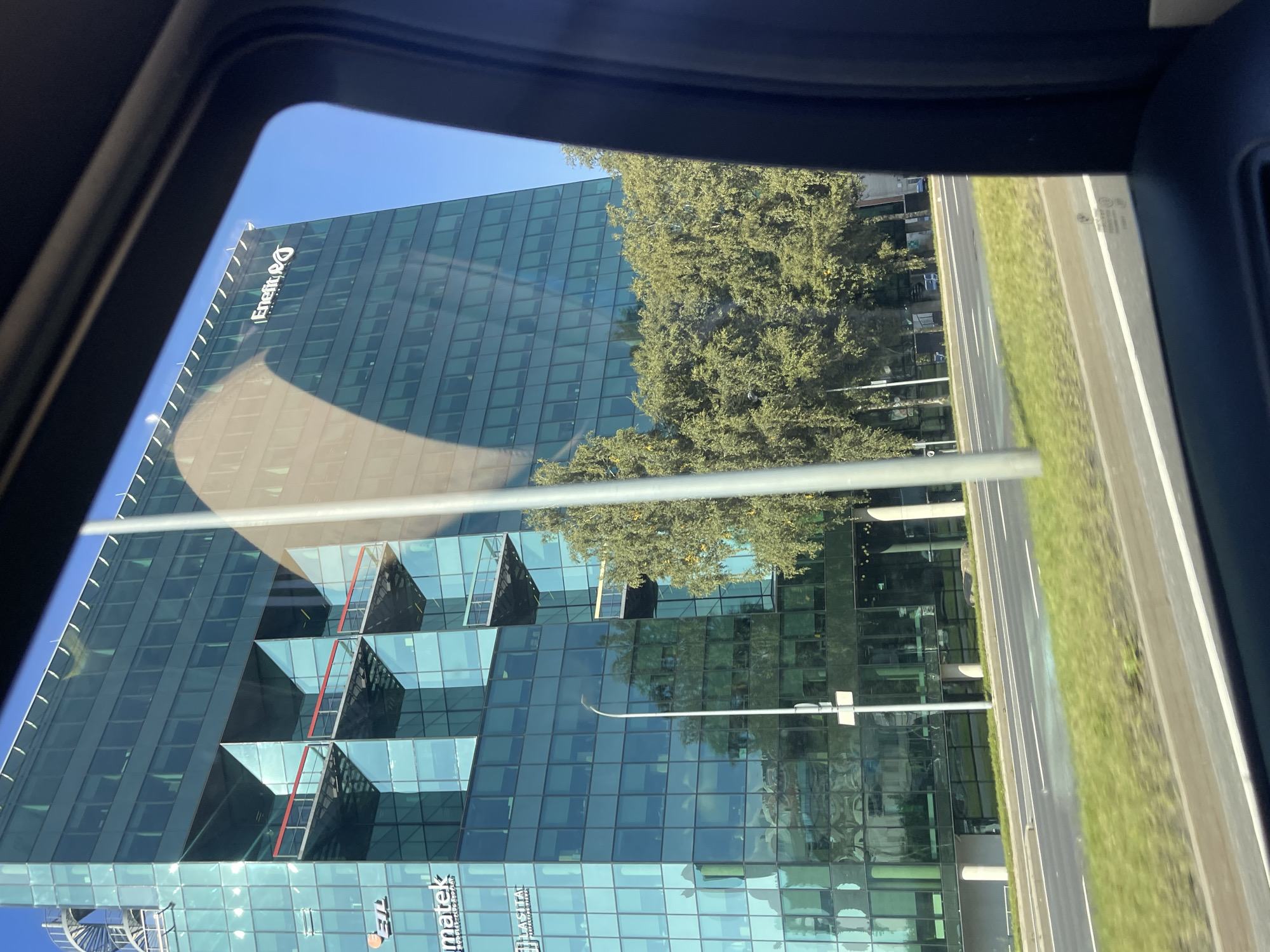 The image shows a modern glass-fronted office building, likely in Tallinn, Estonia, viewed from the interior of a vehicle. The building features a striking architectural design with recessed balconies and is covered in reflective glass panels. Several company logos are visible on the building's facade, including "ETL" and "Enefit". Lush green trees line the street in front of the building, and a road with lane markings is visible in the foreground. The sky is a clear blue, indicating a sunny day. The overall scene depicts an urban environment with contemporary architecture and natural elements.