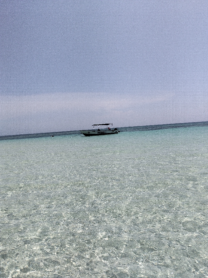 The image depicts a coastal scene in Wasini, Kenya. A single white-hulled, flat-bottomed vessel with a dark canopy is centrally positioned, floating on clear, light turquoise water. The vessel is equipped with two outboard motors at the stern. Legible text on the starboard hull reads 'WASINI 1'. Two indistinct human figures are visible onboard beneath the canopy. A white mooring line extends from the vessel's bow into the water towards the camera's left. The foreground water exhibits exceptional clarity with surface ripples, transitioning to a deeper, darker turquoise towards the distant horizon. The upper frame is occupied by a pale blue-grey sky, appearing hazy or thinly overcast. The scene is illuminated by ambient daylight.