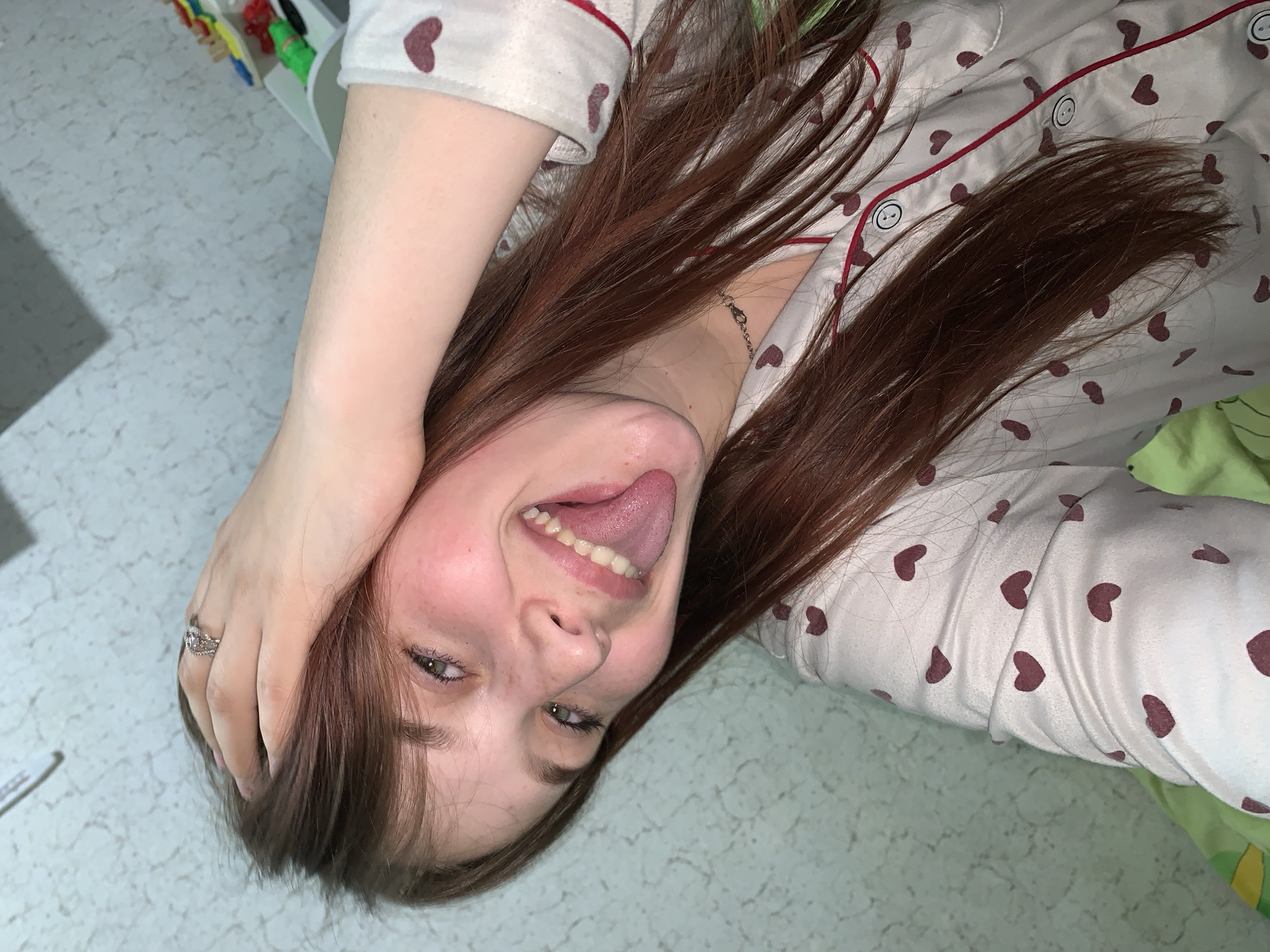 A young woman with brown hair and green eyes is lying down. Her head is tilted to the side, and she is sticking her tongue out. She is wearing a white shirt with red heart patterns. She is wearing a ring on her left hand. The background is a blue-green surface with a textured pattern. The location cannot be determined as Фоки, Russia.