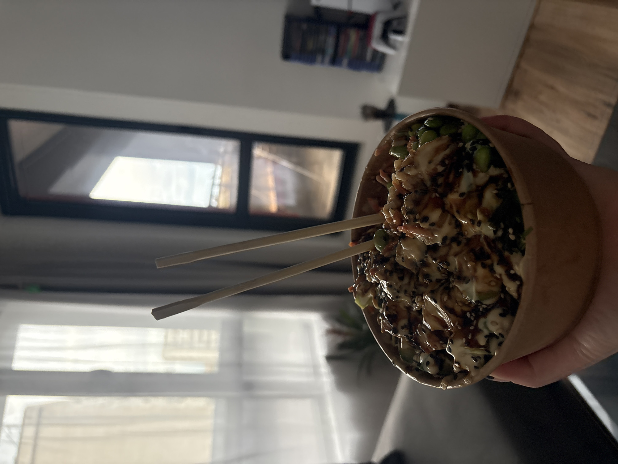 An individual's hand is prominently featured, holding a cylindrical paper or cardboard bowl filled with a meal, likely a poke bowl. The food inside consists of rice as a base, topped with green edamame beans, what appears to be diced protein (possibly chicken or fish), and a generous drizzling of both a creamy white sauce (resembling mayonnaise) and a dark, savory sauce (such as soy or teriyaki). Black and white sesame seeds are scattered over the top, adding texture and garnish. A pair of light-colored wooden chopsticks is inserted into the food, indicating readiness for eating.
The setting is an indoor space, likely a home or apartment in Plovdiv, Bulgaria. In the background, a large window with distinct black framing offers a view of natural light, suggesting it is daytime. Reflections on the windowpanes indicate ambient light conditions, possibly from outside or another room. Above and behind the bowl, shelves stocked with books or other various items are visible, adding to the domestic atmosphere. The lighting is soft and natural, consistent with a daytime indoor scene. There are no explicit emotional cues, specific weather conditions, or any visible text within the frame. The image captures a moment of preparing to enjoy a meal in a comfortable, personal environment.