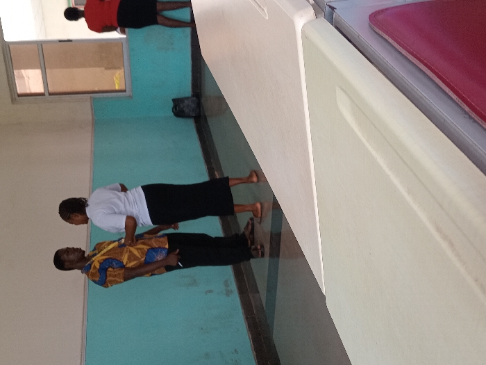 An indoor scene in Ogboko, Nigeria, depicts three individuals in what appears to be a public or institutional building. In the foreground-left, a dark-skinned male, wearing a yellow, brown, and blue patterned short-sleeved shirt, dark trousers, and dark sandals, stands facing a dark-skinned female. He wears a yellow lanyard around his neck and holds a pen in his right hand. The female, barefoot, wears a white short-sleeved top and a long black skirt, with her hair pulled back. They are engaged in direct conversation. In the background-right, another dark-skinned female, barefoot, wearing a red short-sleeved top and a long black skirt, stands facing away from the camera, looking towards a large brown-framed window on a light teal-colored wall. A dark bag rests on the floor near the window. The lower half of the walls are painted light teal, with the upper half cream, separated by a dark trim. The floor consists of dark and light gray tiles. In the immediate foreground, the top surface of a cream-colored counter or bench is visible, partially obscuring the lower half of the scene. A sliver of a red surface is visible beneath the counter on the right.