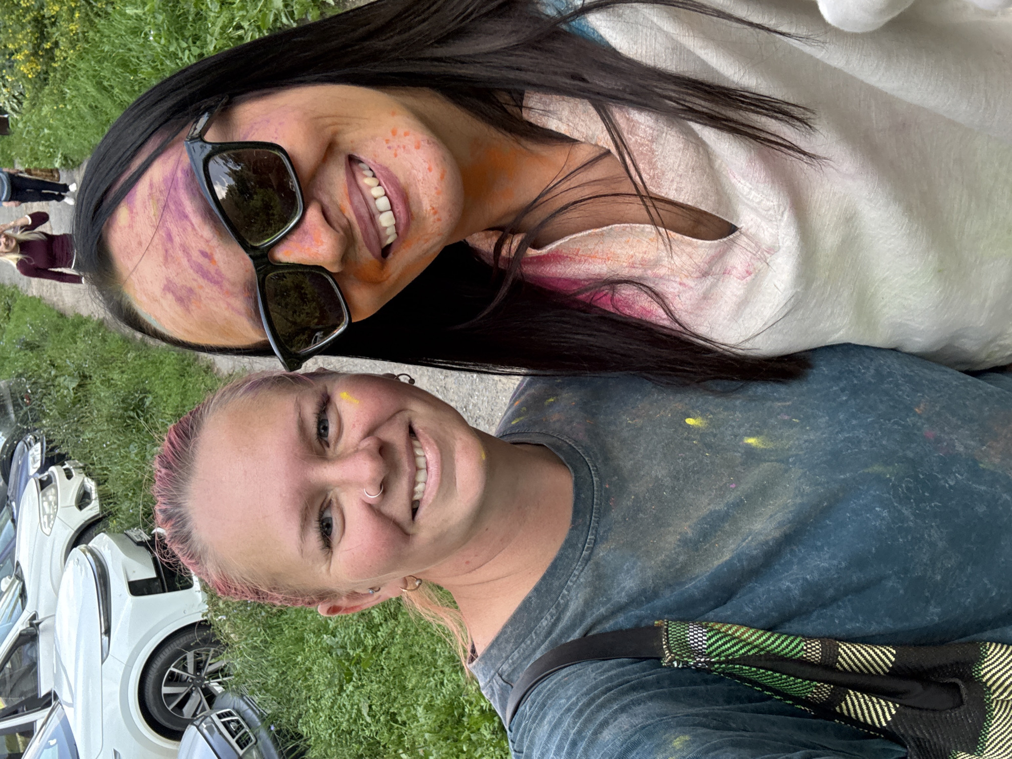 Two women, one with pink hair and the other with dark hair, are posing for a selfie outdoors. Both women have colorful powder on their faces and clothing, suggesting they participated in a color powder event like Holi. The woman with pink hair is wearing a dark teal t-shirt and has a nose ring and hoop earrings. The woman with dark hair is wearing sunglasses and a light-colored top with pink and orange powder on it. In the background, there are several parked cars and lush green foliage, indicating an outdoor setting possibly in a park or a residential area. The presence of cars suggests a location with vehicle access. The overall atmosphere appears festive and casual. The image was taken in Chaikovsky, Russia.
