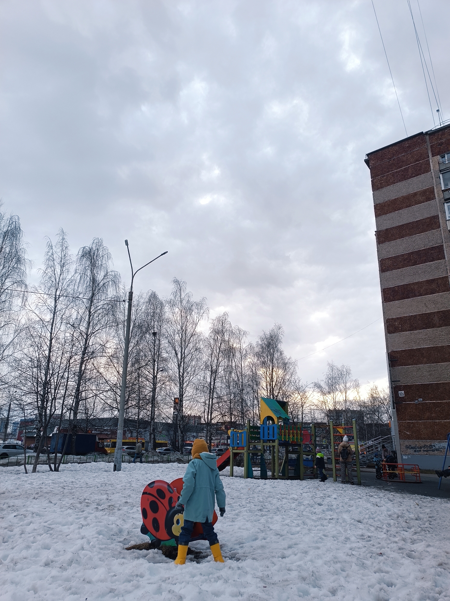 The image depicts a winter scene at a playground in Izhevsk, Russia. A child in a light blue coat, yellow hat, and yellow boots stands in the snow-covered playground near a ladybug-shaped play structure. Other playground equipment, including slides and climbing structures, is visible in the background. Several individuals, possibly parents or caregivers, are present near the playground equipment. Bare trees line the perimeter of the playground. A tall streetlight stands among the trees. In the background, there is a row of buildings and parked cars, indicating an urban environment. A tall apartment building with alternating horizontal stripes of brown and grey is visible on the right side of the image, bearing the address "92a". The sky is overcast with grey clouds.