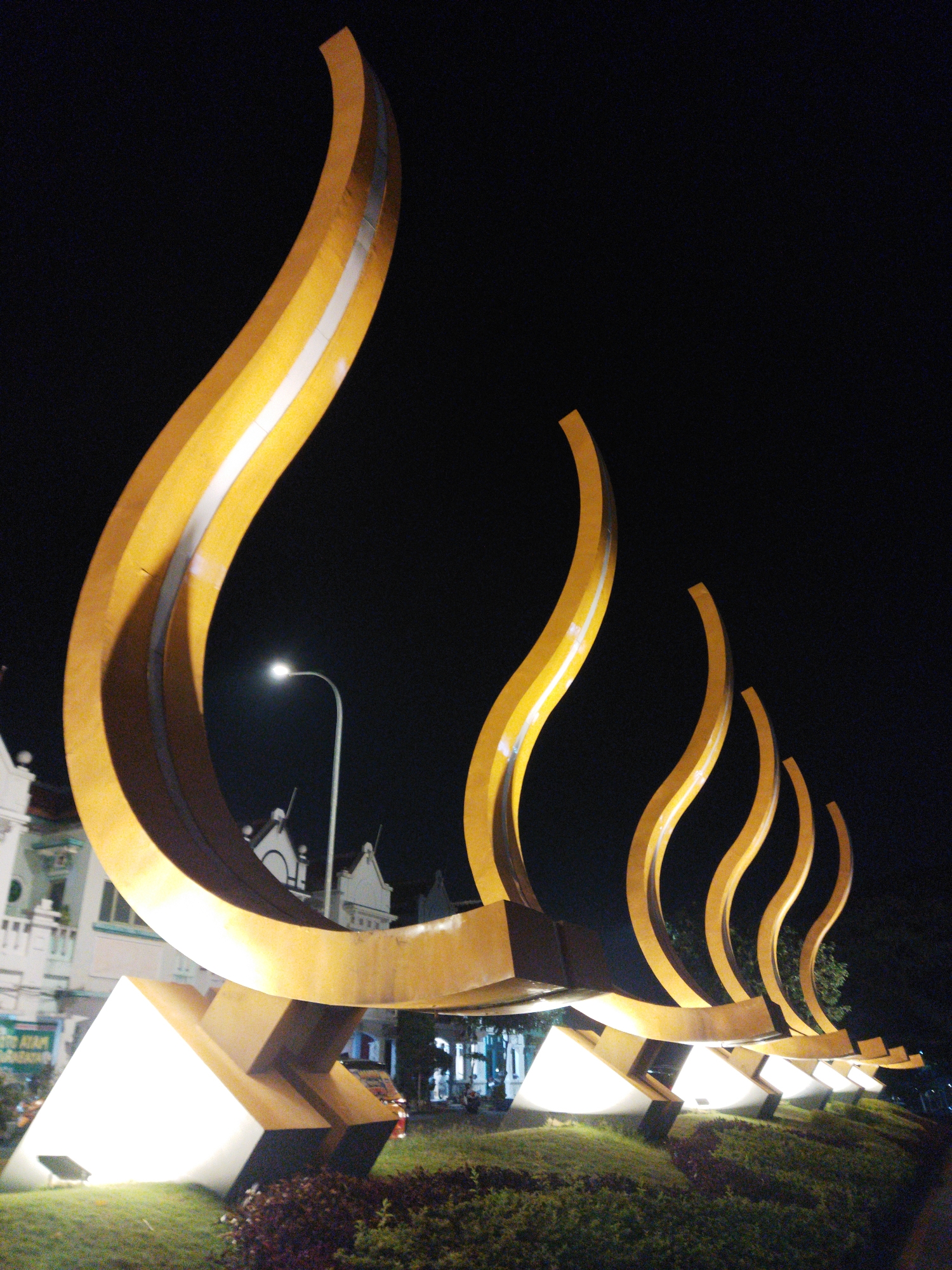 This is a nighttime photograph of a large, modern, golden sculpture situated in Semarang, Indonesia. The sculpture, which appears to be made of metal, features multiple curved, flame-like elements that rise from a horizontal base. The base itself is supported by several illuminated triangular structures, casting a bright glow on the surrounding grassy area. In the background, there are buildings with traditional architecture, a street lamp, and a few trees, all silhouetted against the dark night sky. The sculpture is the primary focus of the image, with its imposing size and striking golden color illuminated by artificial lights.