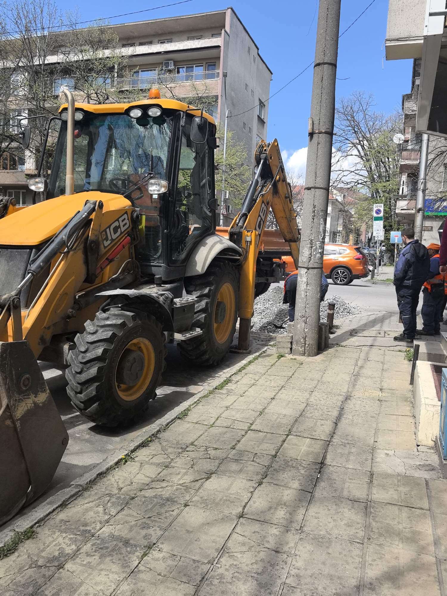 The image captures a street scene in Stara Zagora, Bulgaria, where road repair work is underway. A yellow JCB backhoe loader is parked on the sidewalk, partially blocking it. Construction workers are gathered around a section of the road where gravel is spread. Two workers in safety vests stand on the sidewalk observing the work. In the background, an orange Nissan SUV is parked across the street. A concrete utility pole stands near the construction site. Buildings line the street, and trees with fresh spring foliage are visible. The sky is partly cloudy.