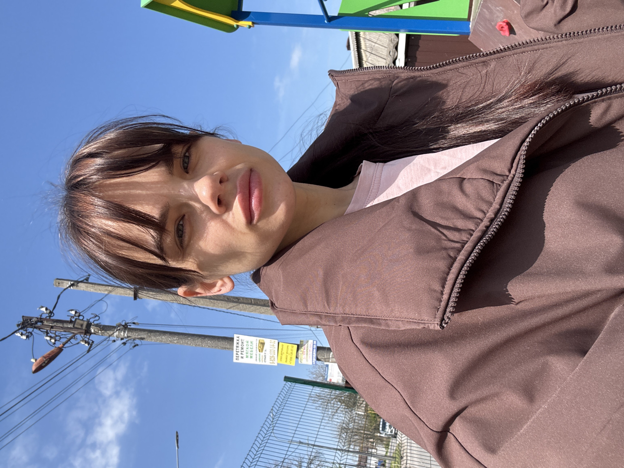 A close-up outdoor selfie captures a young woman looking directly at the camera. She has dark brown hair with bangs and a neutral, slightly pensive expression. Her lips appear full, and her eyes are open. She is wearing a brown zippered jacket over a light pink t-shirt.
The setting is outdoors on a bright, sunny day with a clear blue sky, suggesting morning or afternoon. In the background, to the left, is a concrete utility pole with numerous wires and electrical components. Several signs are attached to the pole; one prominent sign, written in Russian, advertises "ПЕРЕТЯЖКА И РЕМОНТ МЕБЕЛИ" (Upholstery and Furniture Repair) along with a phone number and "СТАМБУЛ A-Я". Another sign nearby says "ИНТЕРНЕТ" (Internet) also with a phone number. A chain-link fence runs horizontally across the middle ground, separating the subject from what appears to be a street or open area. In the upper right corner, hints of brightly colored playground equipment (green, yellow, and blue bars) are visible. The bright sunlight casts distinct shadows, highlighting the textures of her jacket and hair. The overall scene suggests an urban or suburban environment in Rostov-on-Don, Russia.