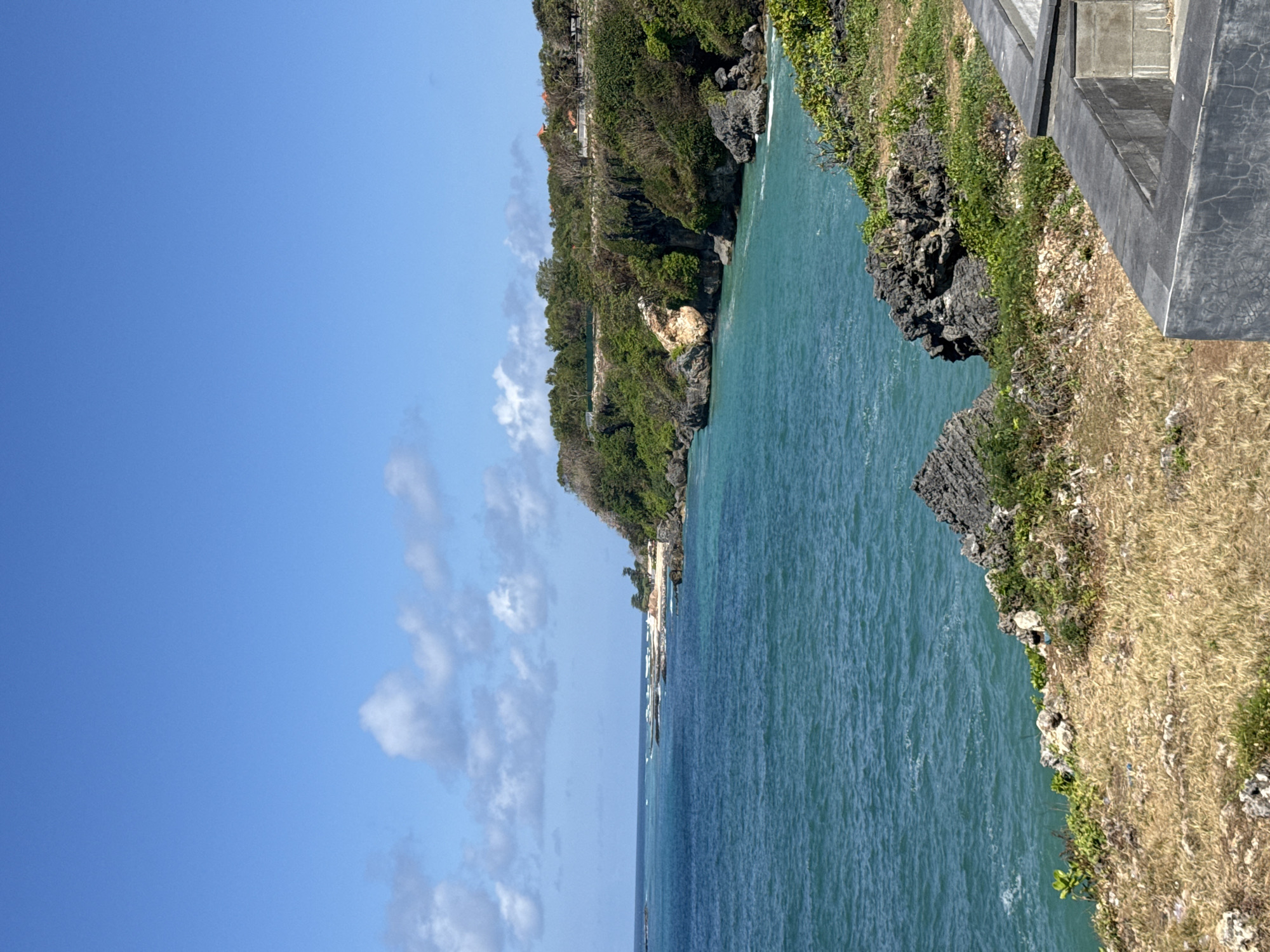 The image depicts a coastal cliffside view in Kuta Selatan, Indonesia. The bright blue sky is dotted with sparse white clouds. Below, a lush green cliff overlooks the turquoise ocean, with rocky outcrops along the coastline. Trees and vegetation cover the cliff face and the top of the plateau. In the foreground, a dry, grassy slope leads down to a constructed grey concrete barrier. The ocean water is a vibrant teal color, with small waves breaking against the rocks. The scene suggests a natural and scenic location, possibly a tourist viewpoint or a natural preserve. There are no people or animals visible in the image.