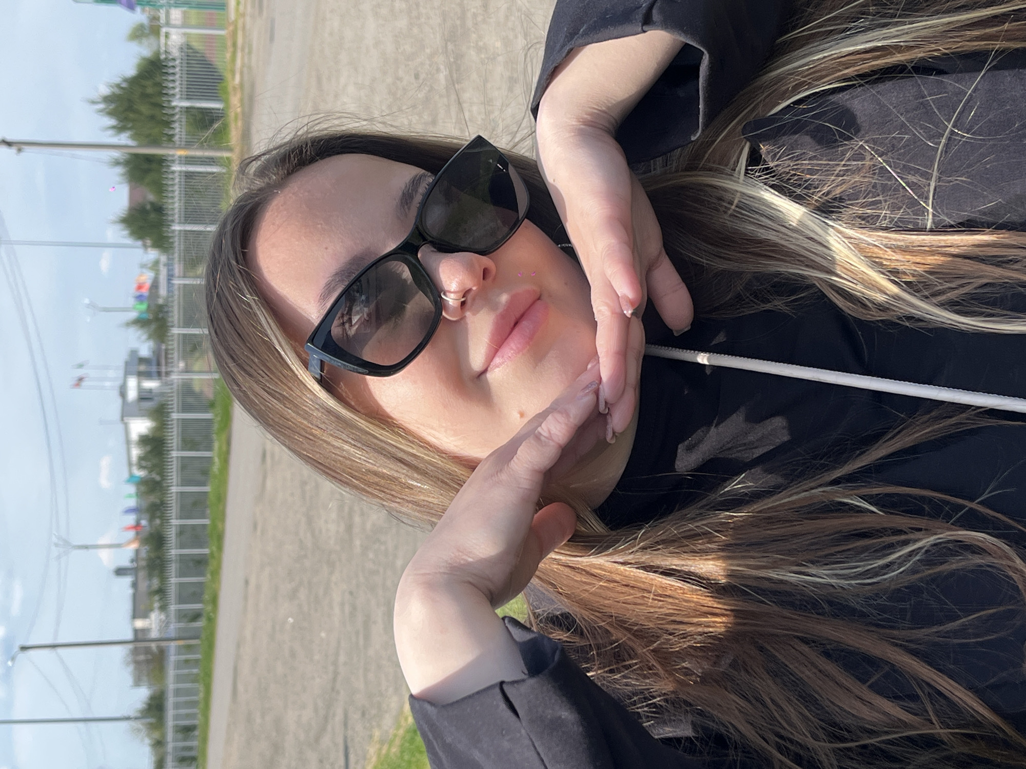 A woman wearing sunglasses and a black hooded top is posing outdoors. She has long, light-brown hair with blonde highlights. The woman's hands are positioned around her face. A silver ring is visible in her nose. The background shows a grey paved area, grass, a fence, and distant buildings, indicating an urban or suburban setting. In the background are colored flags. Considering the location of Novopokrovskaya, Russia, the scene is likely situated in an outdoor area, possibly a park or open space within the city.