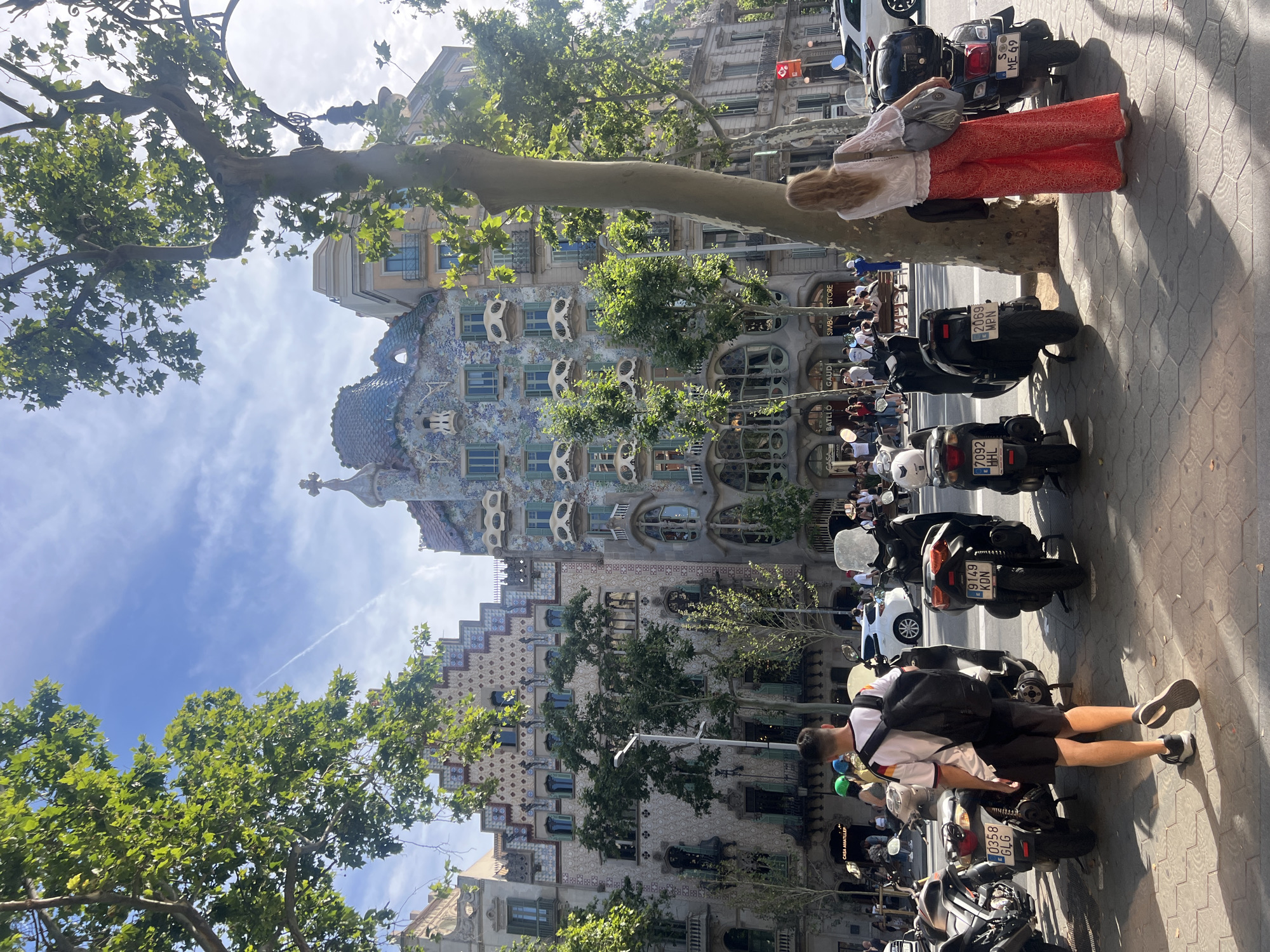 In Barcelona, Spain, on a sunny day, a man wearing a white t-shirt with black and yellow stripes, black shorts, and a black backpack walks away from the camera on a sidewalk. He is walking past several parked scooters, some with passengers. To the right, a woman with long blonde hair, wearing a white blouse and red patterned pants, stands near a tree, looking at the buildings. In the background, the ornate facade of Casa Batlló, a famous modernist building designed by Antoni Gaudí, is visible. The building features colorful mosaics, sculpted balconies resembling bone structures, and a distinctive roof. Trees line the sidewalk, casting shadows on the ground. The sky is blue with some wispy clouds.