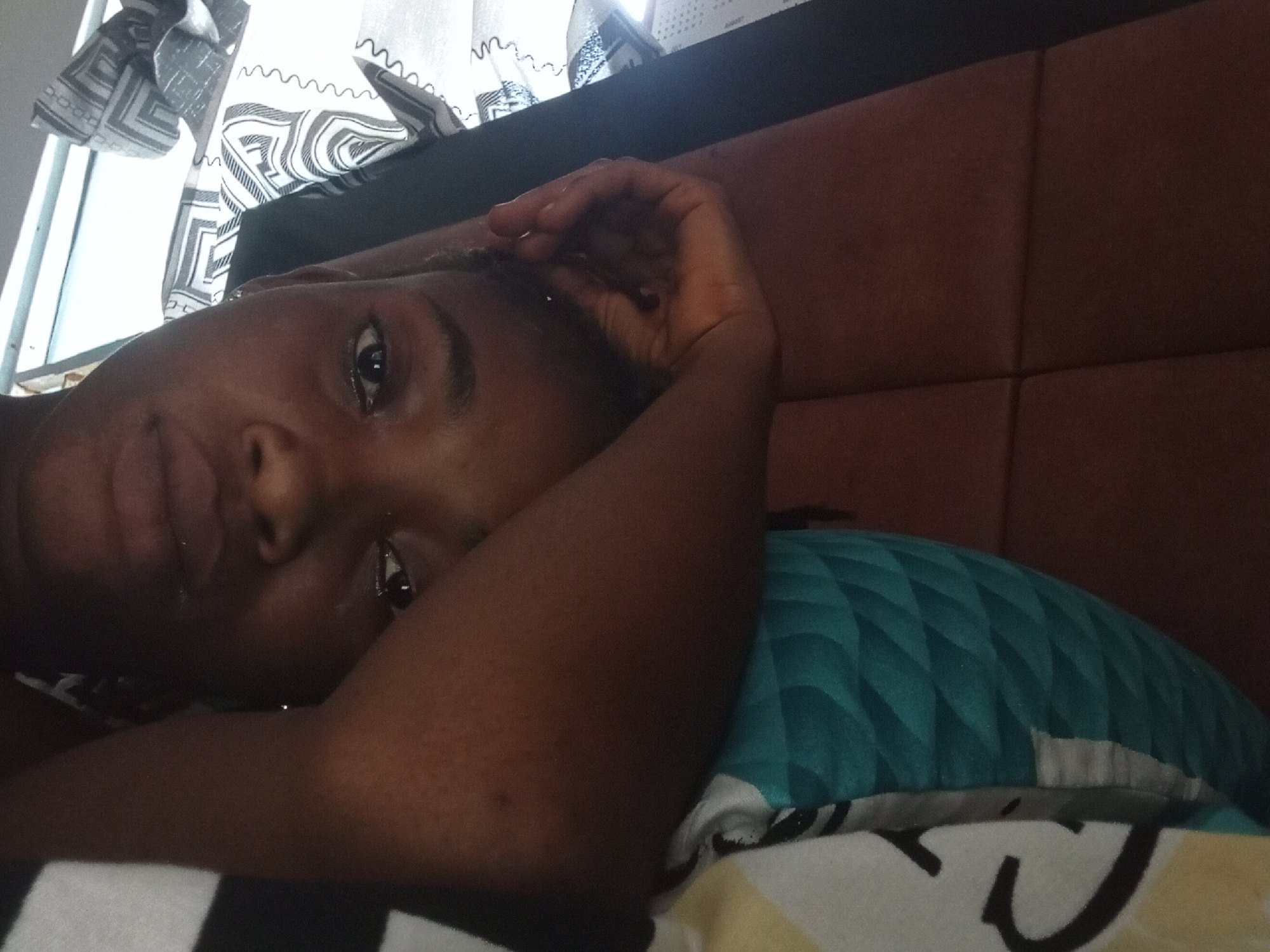 A close-up indoor shot features a dark-skinned person lying on their side. They are resting on a pillow with a teal and white geometric pattern, positioned against a brown upholstered headboard. The person's left arm is bent, with their hand near their forehead. The individual's face is turned towards the camera with a direct gaze. A window is visible in the upper-left corner with white curtains featuring a black and white geometric design. The location is not specified.