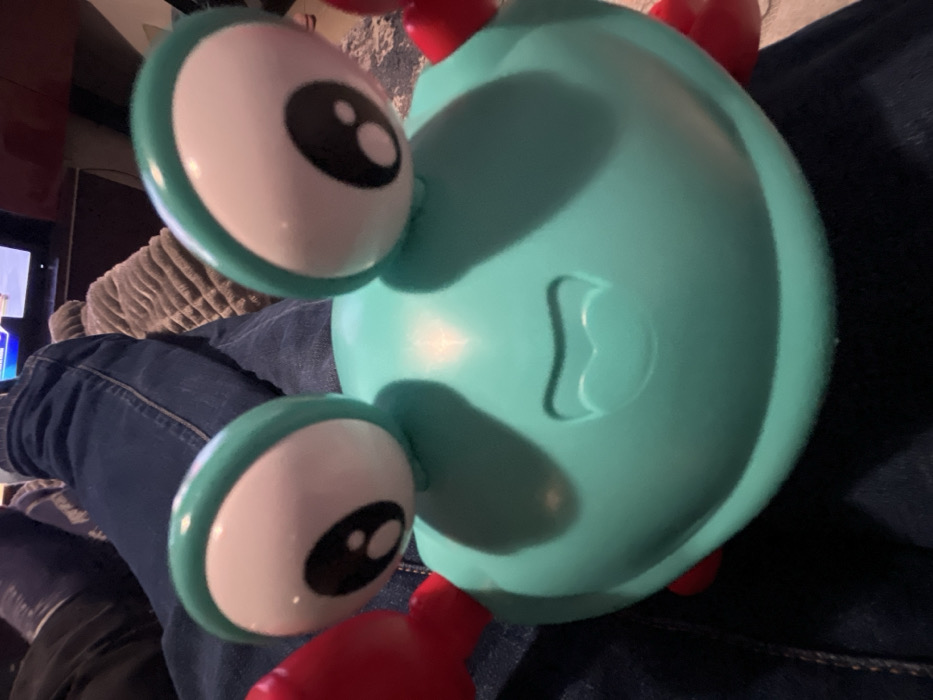 A teal toy crab with large, white eyes with black pupils rests on a lap clad in blue jeans. The crab has red legs and a semi-circular body with a molded shape near the center. The background includes a couch and a television in a dimly lit room.