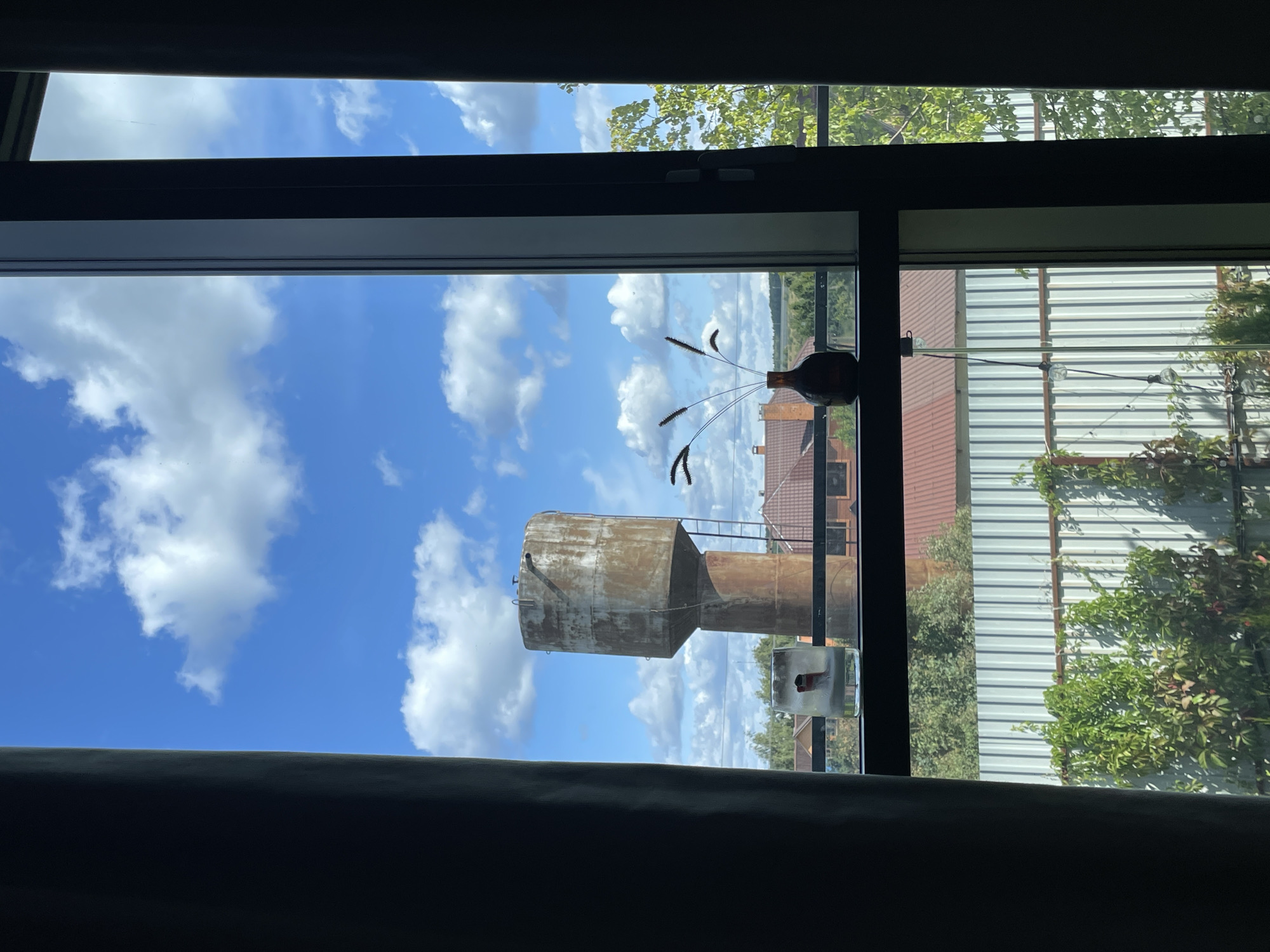 The image shows a view through a window in Pokrovskoe, Russia. Outside, a large, weathered concrete water tower stands prominently against a bright blue sky with scattered white clouds. Below the tower, residential rooftops with red tile are visible, along with a tall, corrugated metal fence and lush green foliage. On the windowsill inside, two decorative items are placed: a glass with a red and white figure and a small brown vase holding dried grass-like stalks. The scene is framed by dark curtains on the left and right sides of the window.