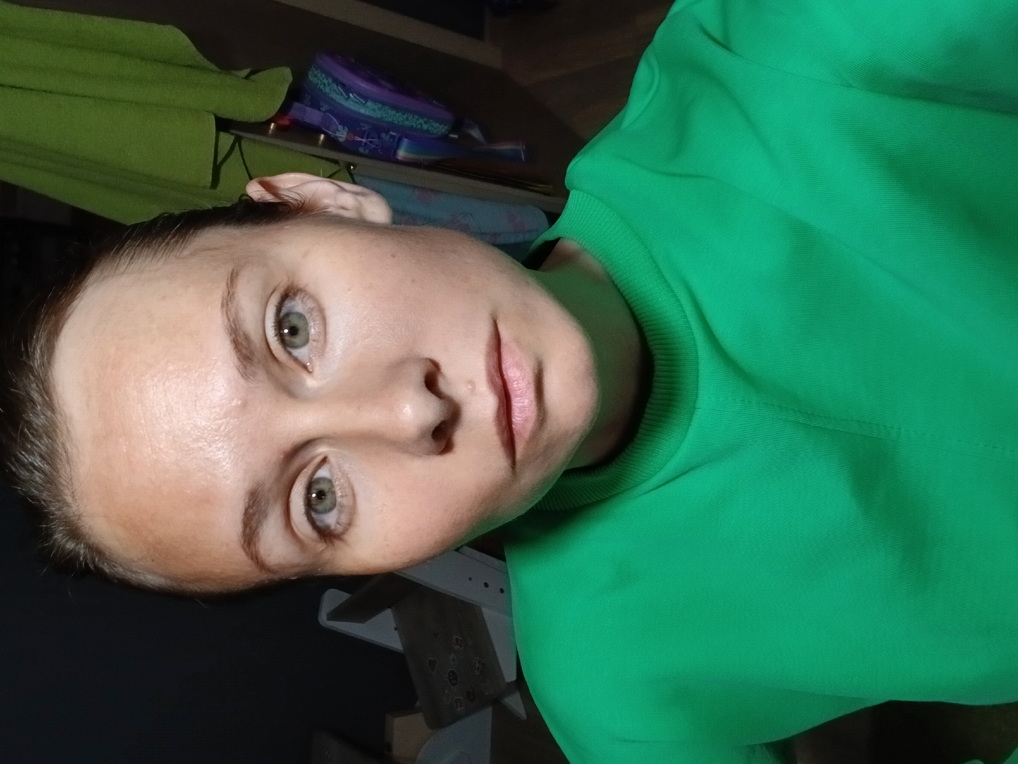 A woman with light skin and short brown hair pulled back is facing the camera, with her head slightly tilted. She has light green eyes and appears to be wearing light makeup. She is wearing a bright green crew-neck sweatshirt. The background is dark, with some objects visible including a green draped fabric on the right and what appears to be furniture and a backpack on the left. The image was taken indoors.