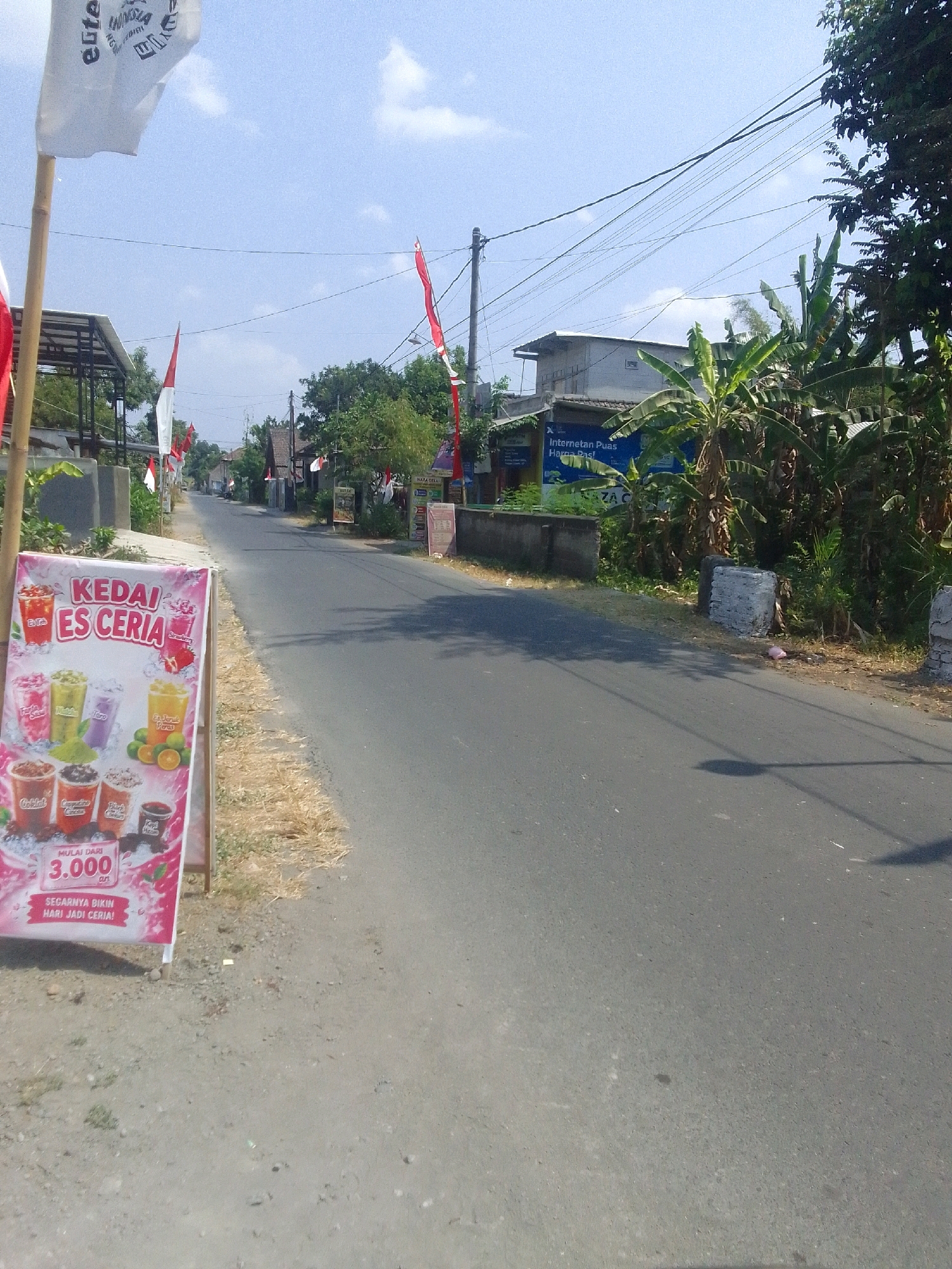 The image depicts a street scene in Ngadiluwih, Indonesia. A white banner with red Indonesian flags is visible on the left side of the image, near a bamboo pole. In the foreground, a pink and white advertising sign for "KEDAI ES CERIA" (Cheerful Ice Shop) displays various beverages and a price starting from 3.000. The sign also includes the tagline "Segarnya bikin hari jadi ceria!" (Its freshness makes the day cheerful!). The street is paved with asphalt and extends into the distance, lined with trees and buildings. Overhead, power lines are strung across the sky. In the middle ground, there's a building with a blue advertisement that reads "Internetan Puas, Harga Pasti!" (Satisfying internet, Sure Price!). A few red and white flags are also seen planted along the side of the road. The sky is clear and blue with scattered clouds.