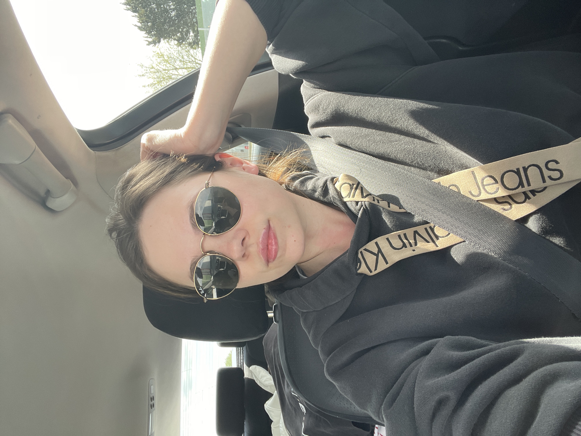 The image depicts a woman wearing sunglasses and a black Calvin Klein hoodie, seated in a car. She is wearing a seatbelt. The interior of the car is visible, with the sunroof open. The location is Vitulazio, Italy, as indicated in the prompt. The woman is holding her hand on her head. Outside the car, trees are visible.