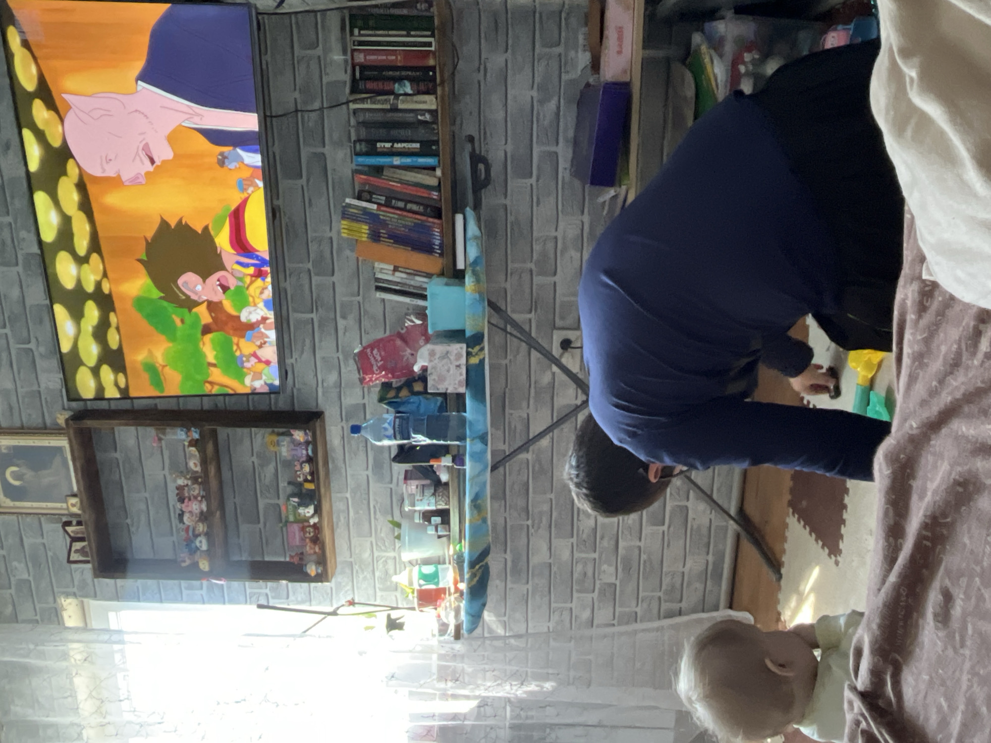 An indoor scene captured from a slightly elevated, rotated perspective shows two individuals in a room with faux brick wallpaper. A man, wearing a dark blue long-sleeved shirt and black pants, is bent over, facing away from the camera, seemingly engaged with something on the floor, possibly toys (yellow and green ones are visible). To his right, a baby with blonde hair and a light-colored top sits on a light-brown patterned blanket or rug, looking towards the floor or the man.
In the background, a television screen displays an animated show, possibly an anime like Dragon Ball, featuring characters including a pink-skinned, pig-like figure and a brown-haired figure with spiky hair. Below the TV, a dark wooden shelf unit holds various small figurines or toys. To the left of the TV, another wooden shelf is filled with numerous books, some with titles clearly visible in Cyrillic script, such as "ОТЕЦ ААРСЕЙ" (Father Arseny), and partially readable titles like "КАК НАЧАТЬ ЗАНИМАТЬСЯ" (How to Start Doing) and "ВЕРНЕМСЯ НА РОДИНУ" (Let's Return to the Motherland), indicating a Russian context.
A light-colored folding table, possibly used as a desk or work surface, stands between the man and the bookshelves, holding a water bottle, various containers, and other small items. Bright light streams into the room from a window on the left. The floor appears to be a mix of wood and soft, interlocking foam mats in brown and tan hues. A framed picture of a black cat looking at a crescent moon hangs on the wall to the left of the shelves. The time of day appears to be daytime due to the natural light.