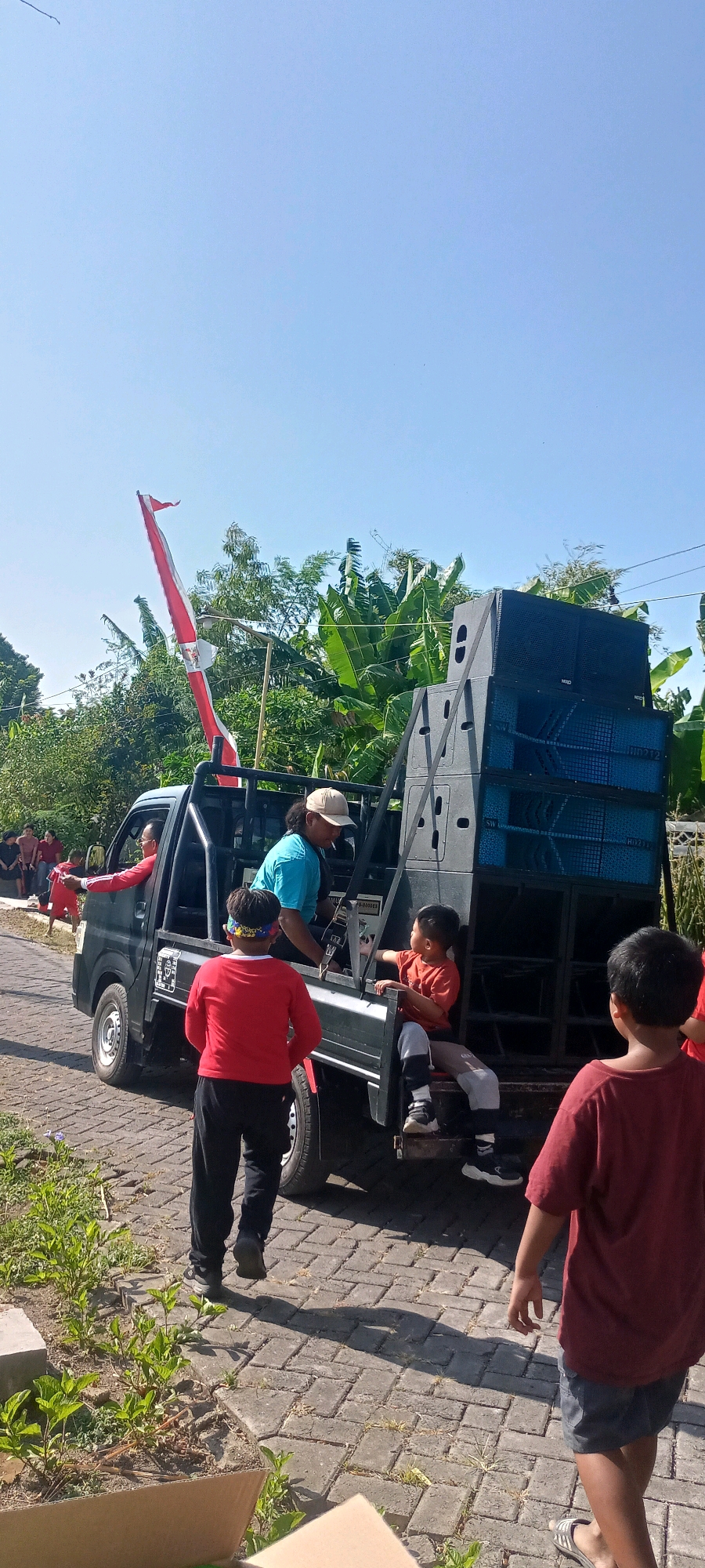 In Gempolklutuk, Indonesia, on a sunny day with a clear blue sky, a small black pickup truck is parked on a paved road. The truck is loaded with a large stack of dark blue and gray speakers. Several people are gathered around and in the truck. In the driver's seat, a man wearing a red shirt is visible. In the back of the truck, a man in a blue shirt is seated and smoking a cigarette. Two young boys are also in the truck bed. One boy, wearing an orange shirt, is sitting and appears to be interacting with the man in the blue shirt. Another boy, wearing a red shirt and a patterned headband, is standing. Further back, a boy in a maroon shirt and dark shorts is standing facing away from the camera. A red and white flag is prominently displayed on a pole attached to the truck. The surrounding environment includes green trees and foliage.