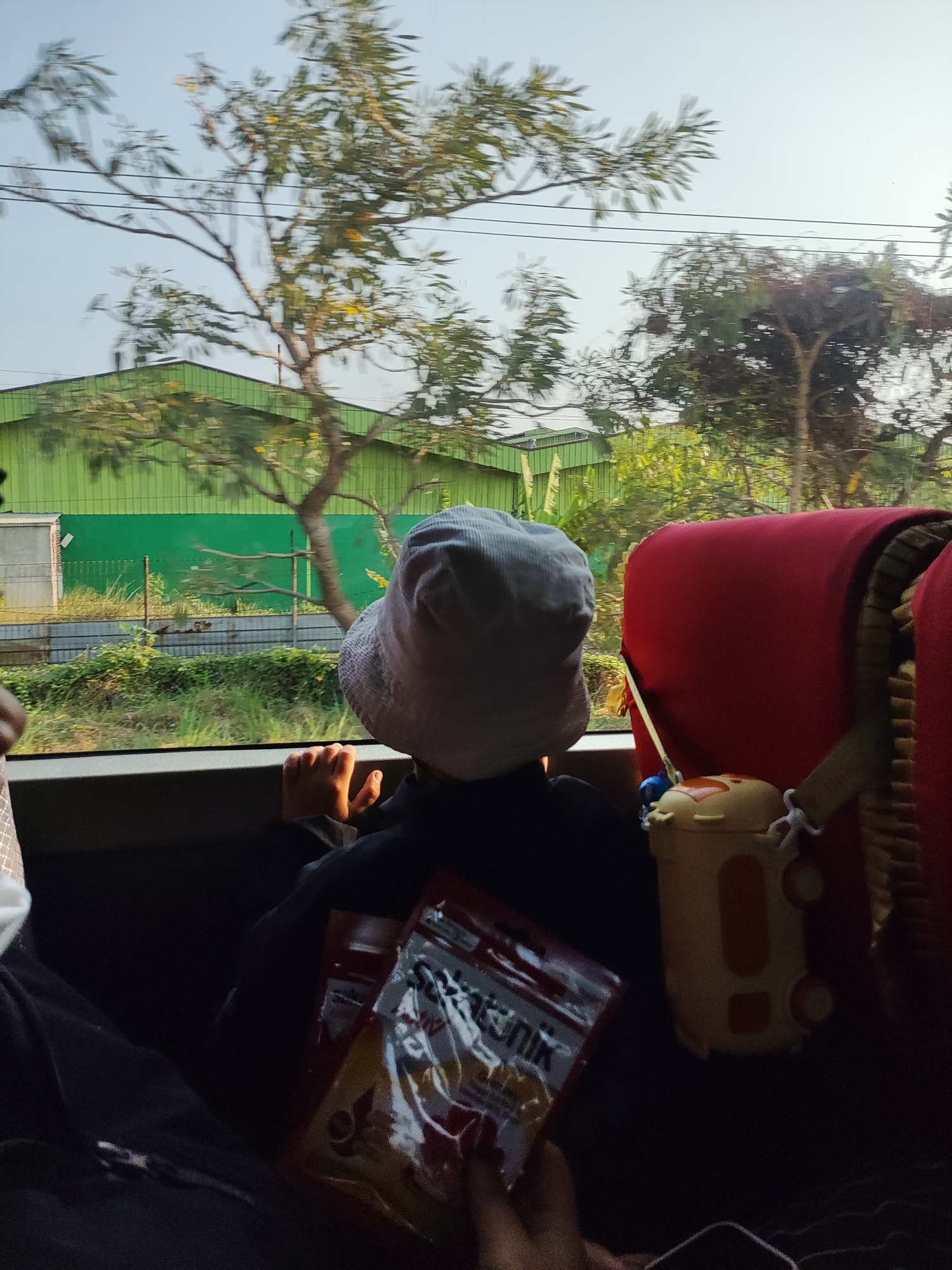 The image shows a child wearing a white bucket hat and a dark jacket, sitting in a vehicle and looking out the window. The child's hands are resting on the window sill. Outside the window, a green building with a corrugated metal facade is visible, along with trees and vegetation. A fence separates the area outside the vehicle from the road. A yellow and orange toy, possibly a lunchbox or dispenser, is attached to the back of a red seat in the foreground. A person's hand, holding a packet of snacks, is visible in the lower right of the frame. The scene suggests a journey, possibly through the outskirts of Karawang, Indonesia, characterized by industrial buildings and natural landscapes.